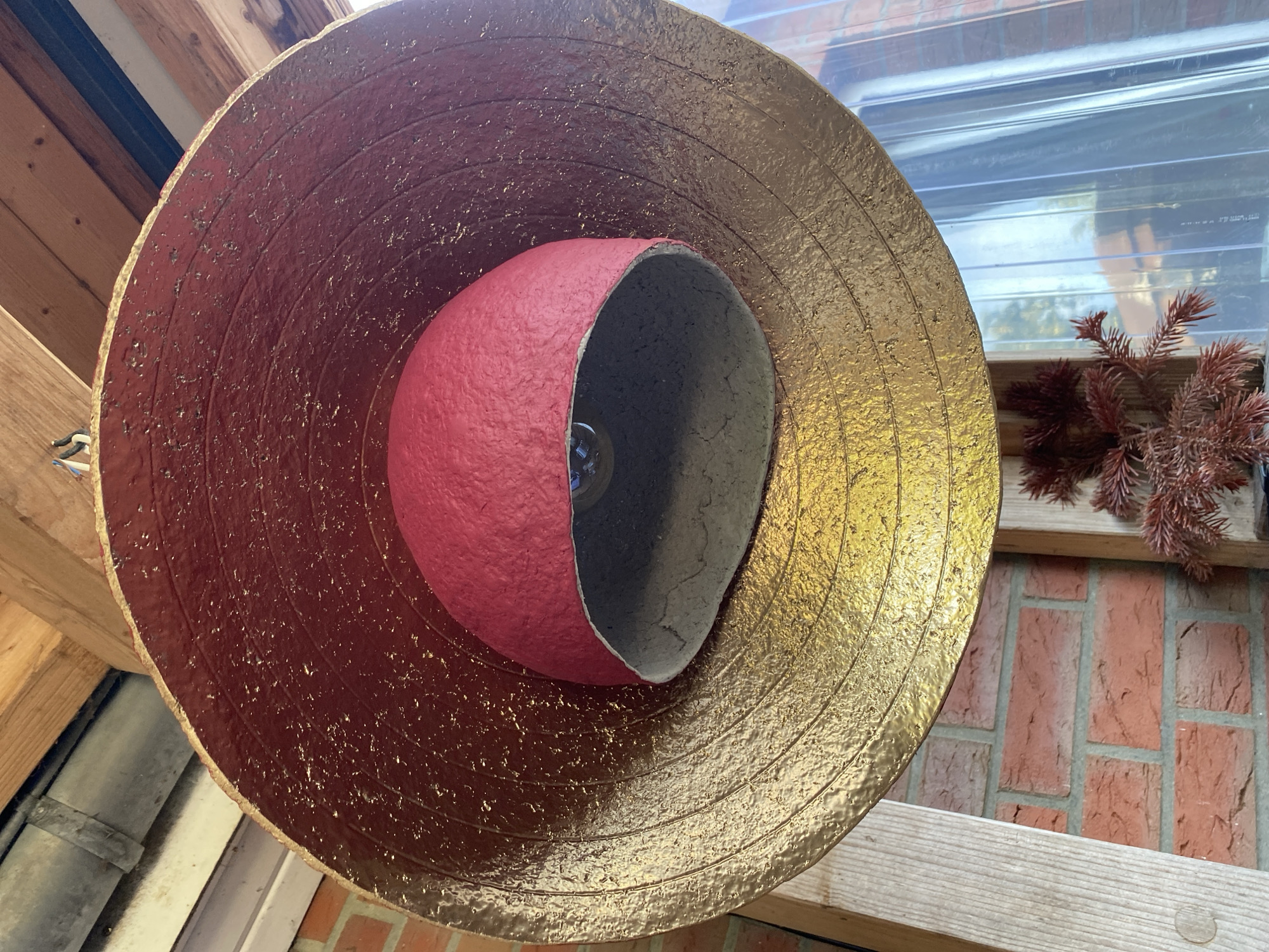 An upside-down, handmade lampshade hangs from wooden beams. The lampshade is conical with concentric rings pressed into its surface and is painted a metallic gold. Inside, a smaller, spherical shade, painted red, is suspended. A clear lightbulb is visible within the inner shade. The background shows a brick wall, a clear plastic sheet, and some dried foliage. The image was taken in Lütjenwestedt, Germany.