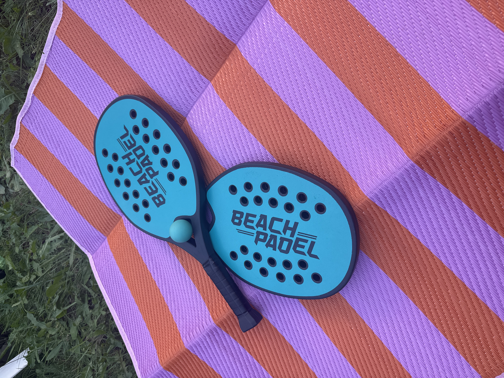 Two turquoise beach paddles and a small turquoise ball are laid out on a striped mat. The mat has alternating wide stripes of orange and lavender. The paddles are positioned diagonally on the mat, with one overlapping the other. The top paddle has the words "BEACH PADEL" printed in black on its surface, and it also has a small turquoise ball resting on it. The bottom paddle also has the words "BEACH PADEL" printed in black. The mat is placed on a grassy area, likely outdoors. There are no people visible in the image. The scene appears to be set in Katowice, Poland, suggesting a recreational or park setting.