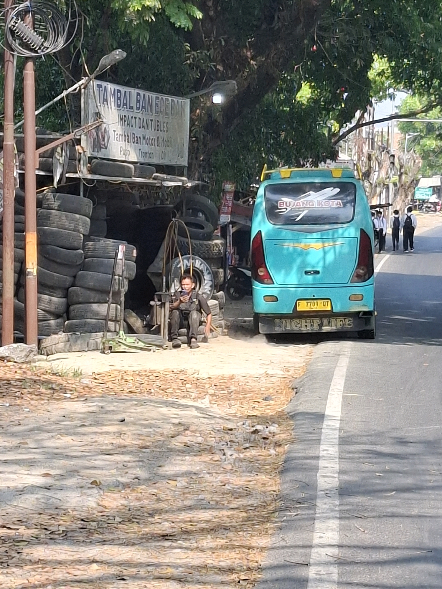 The image depicts a tire repair shop in Sukabumi, Indonesia, a city known for its natural beauty and agricultural products. The shop, located roadside, is characterized by stacks of used tires forming the structure of the establishment. A sign above the entrance reads "TAMBAL BAN ECE DAN IMPACT DAN TUBLES Tambal Ban Motor & Mobil Pusp Tronton DD". A man is seated on a chair inside the shop, looking at his phone, surrounded by tires. In the background, a teal-colored bus with the words "BUJANG KOTA" and an airplane graphic on its rear window is parked on the street. The bus has a license plate that reads "F 7709 QT" and the words "NIGHT LIFE" are visible on its bumper. Several individuals, likely customers or passersby, are walking on the sidewalk in the distance. The environment is sunny with shadows cast by trees, suggesting a warm climate. The road is paved, with a white line marking the lane. The ground in front of the shop is dirt and gravel, with scattered leaves. The overall scene conveys a typical street-side business operation in a developing urban area.