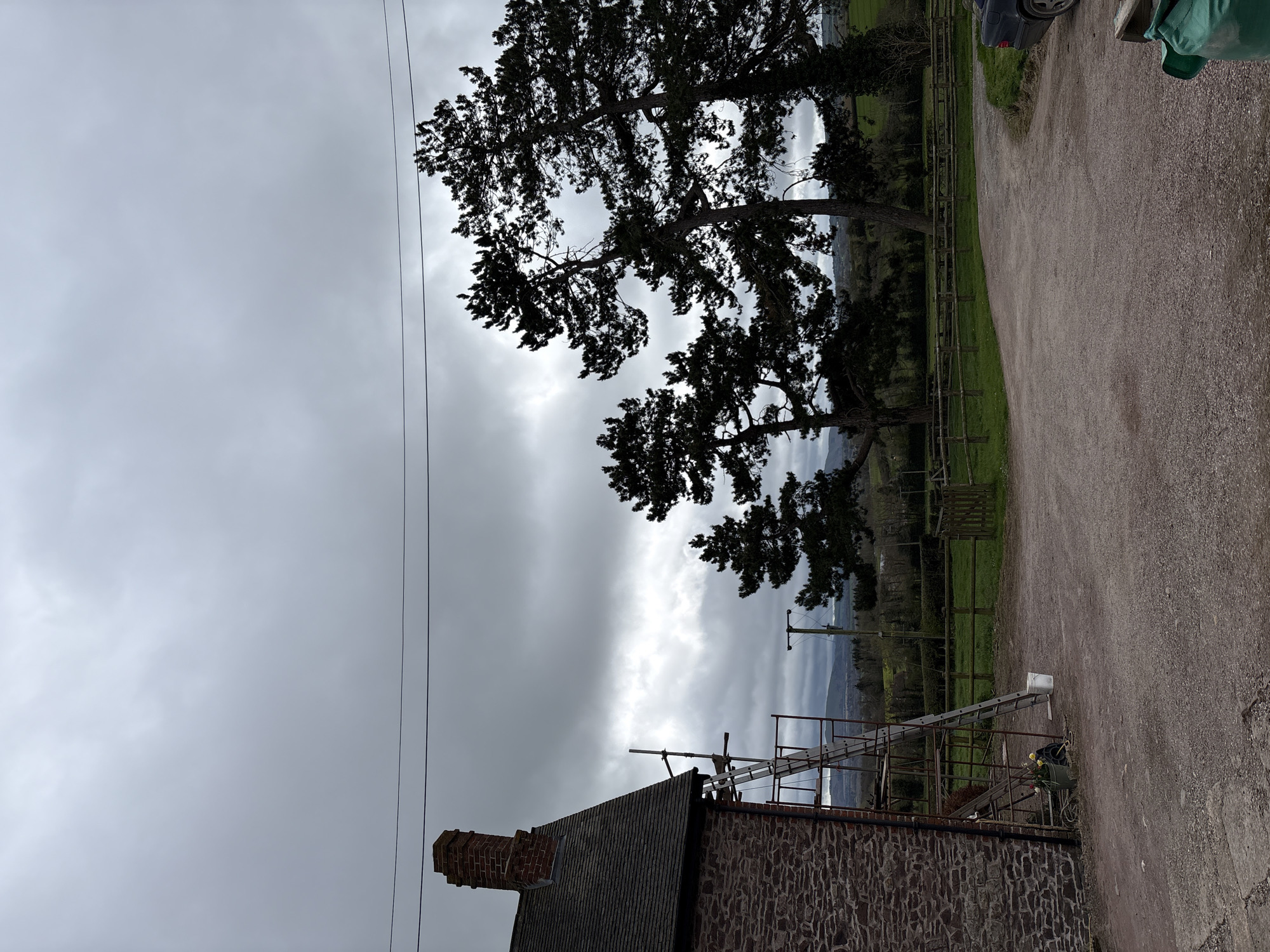 The image depicts a rural scene in Longden, United Kingdom. A stone building with a chimney occupies the foreground, its roof partially covered with scaffolding and a ladder. Overhead power lines are visible against a cloudy sky. In the background, silhouetted trees and a distant landscape with fields, fences, and additional power poles are discernable. A gravel driveway is to the right. A car is partially visible.