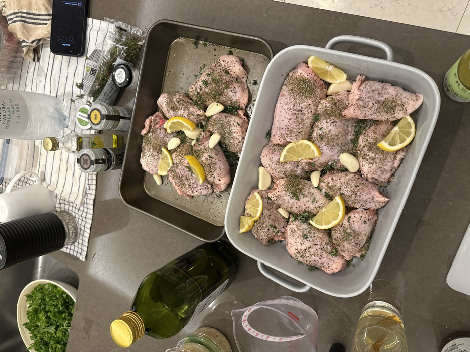 Two pans of seasoned chicken with lemon slices and garlic cloves sit on a countertop. Spices, herbs, and olive oil bottles are scattered nearby. A bowl of chopped greens, a water pitcher, and a phone are visible on a striped towel. A measuring cup and a glass with a lemon slice are also on the counter.