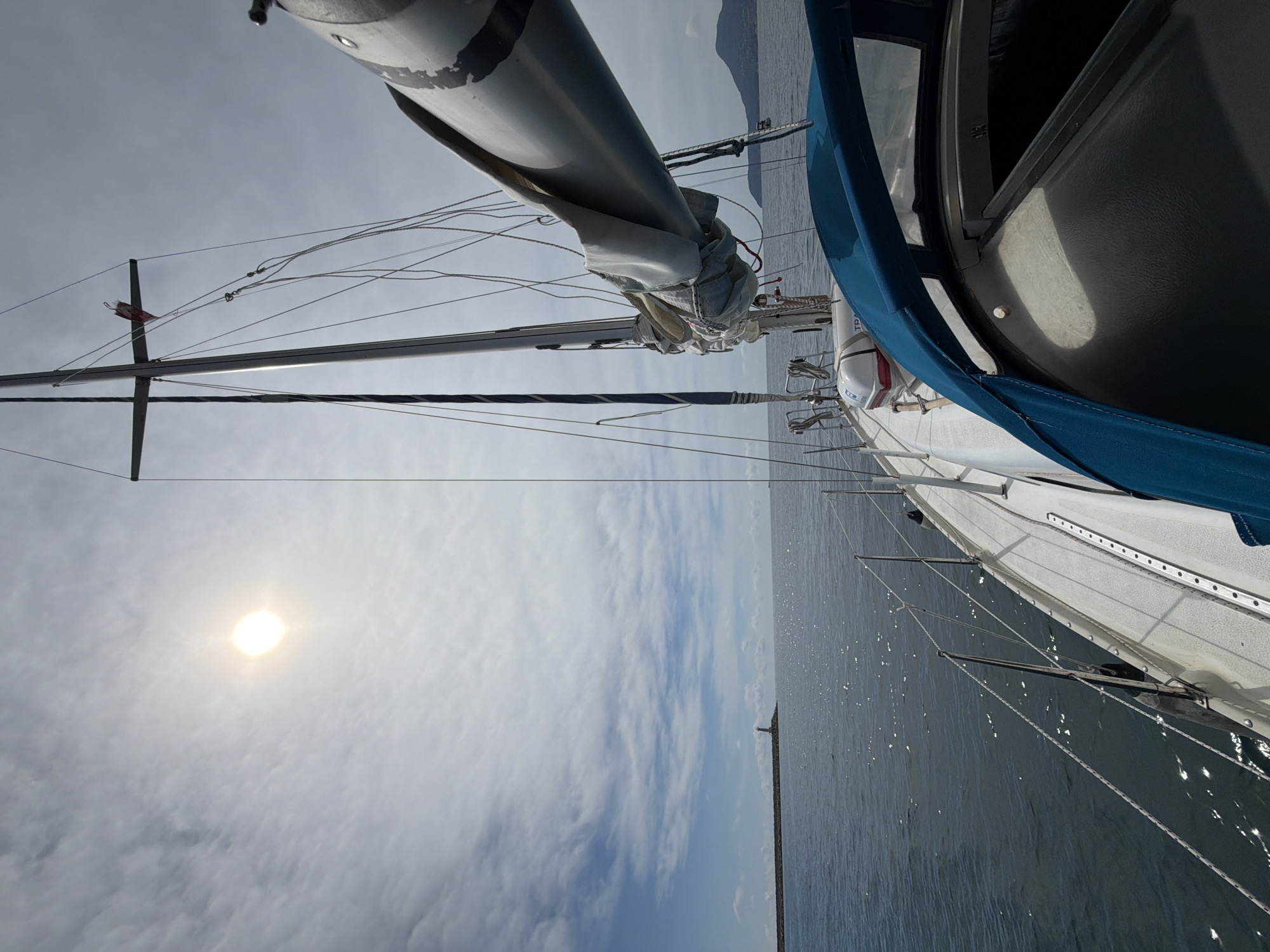 This is a vertical, wide-angle, eye-level shot taken from a sailboat, likely in the Bar, Montenegro area. The camera is positioned on the deck, looking forward and slightly upwards. The main subjects are the sailboat itself, with its mast, rigging, and furled sail prominently featured against a cloudy sky. The sea, with its choppy surface reflecting sunlight, occupies the right side of the frame. In the distance, a long breakwater with a lighthouse or small structure is visible.
The activity is sailing on a bright, though partly cloudy, day. The sun is visible, indicating it's likely daytime, perhaps late morning or early afternoon. The weather appears fair, with scattered clouds and bright sunshine. The presence of the sea and the sailboat clearly places the scene in a maritime environment.
Notable details include the intricate network of ropes and wires supporting the mast, the blue canvas covering part of the boat's structure, and the textured surface of the deck. The water is a dark blue-grey, with many small, bright reflections indicating movement and light. The overall mood is peaceful and scenic, capturing a moment of sailing. There is no discernible text in the image.