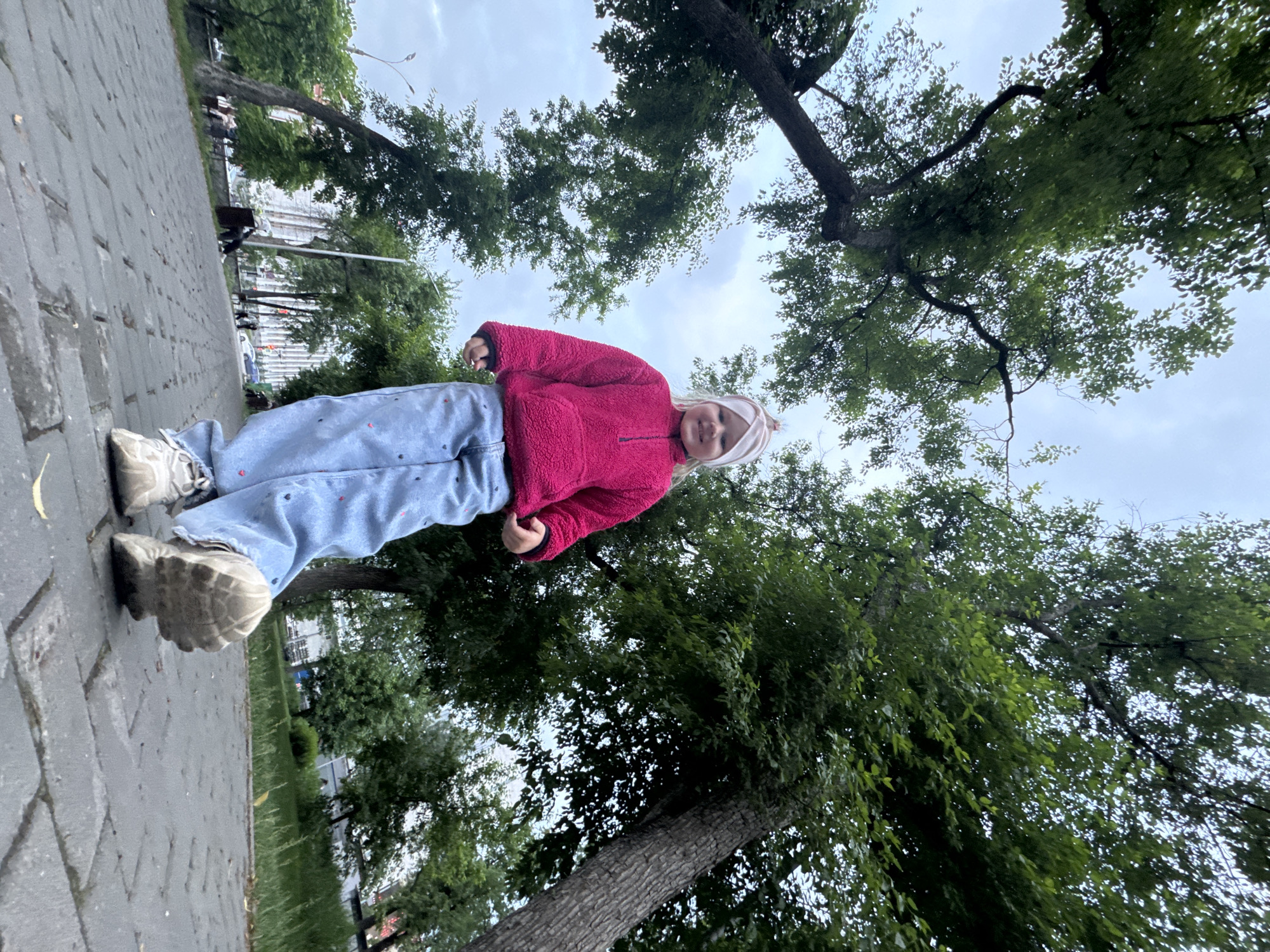 A photograph taken at an odd angle depicts a young child standing on a paved walkway. The location is the city of Perm, Russia, as identifiable from the city's characteristic architecture in the background and vegetation. The child, wearing a pink fleece jacket, printed jeans, and light-colored sneakers, looks up towards the camera. Tall trees frame the sky, providing a backdrop of green foliage and an overcast sky. The paving stones underfoot suggest a park or pedestrian area.