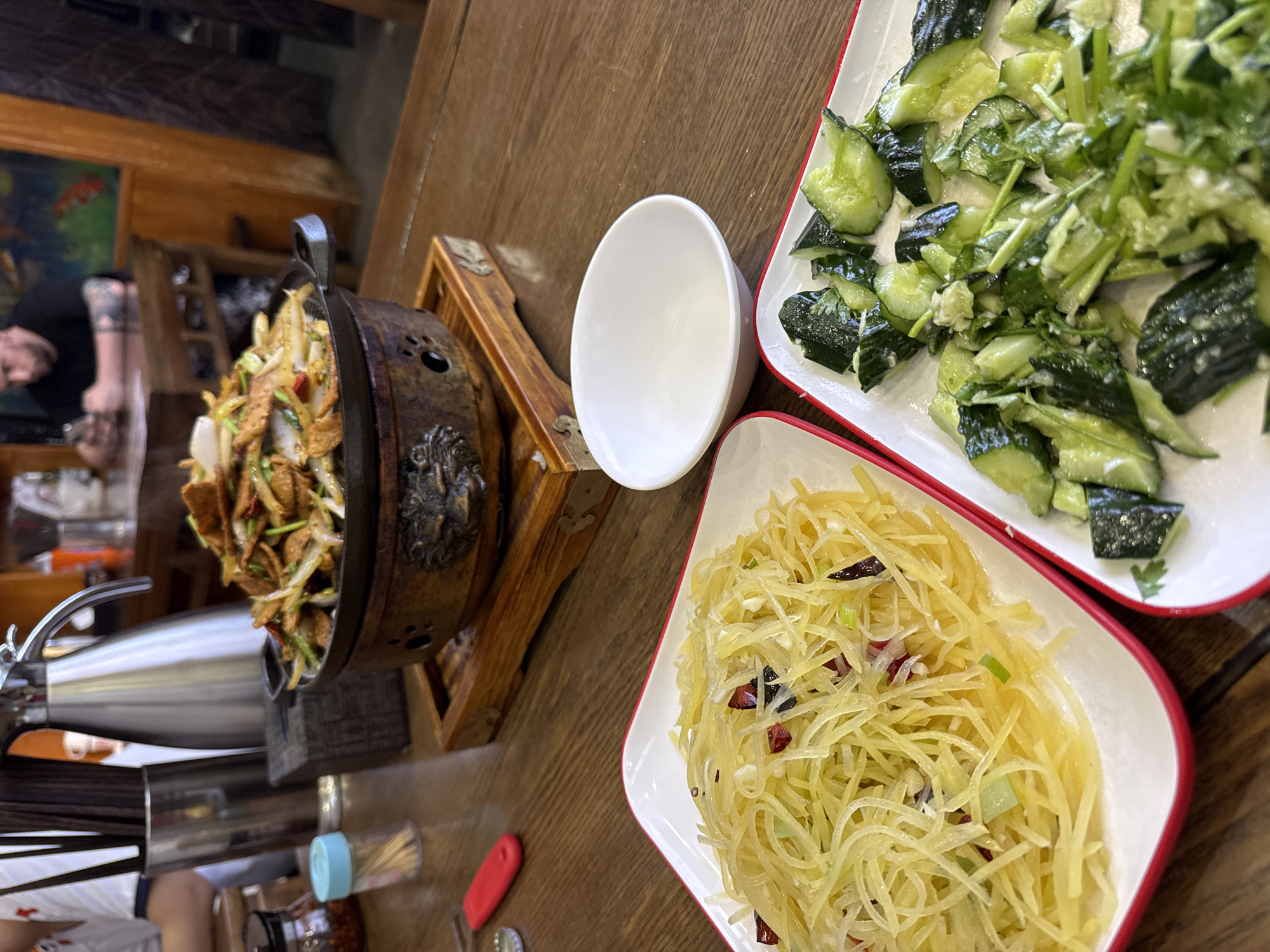 On a wooden table, presumably in a restaurant in the Dongcheng District, China, are several food items. In the center-left, a dark metal pot with a handle rests on a wooden stand, filled with a stir-fry-like dish with greens and browned pieces. To the right, two white plates with red rims contain cold dishes: a plate of cucumber salad and a plate of shredded potatoes mixed with red chilies. Beside the pot is a small white bowl, a metal teapot, and condiments. A person can be seen in the blurry background, suggesting an indoor setting.