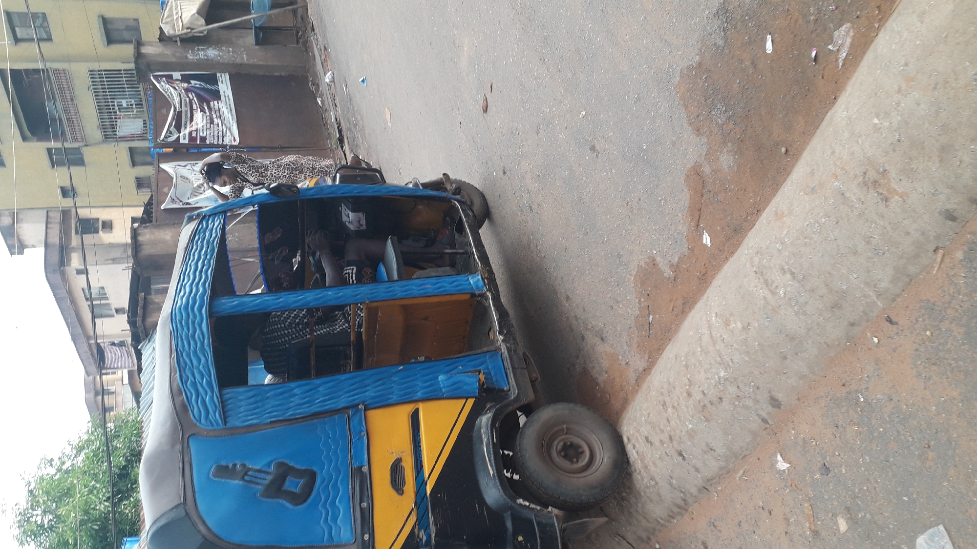 The image shows a blue and yellow commercial tricycle, locally known as a "Keke," parked on the side of a street in Onitsha, Nigeria. A woman in a patterned dress leans into the open passenger area of the Keke. Other passengers are visible inside. Buildings are in the background, including one with signage. The street surface appears worn, with some discoloration and debris. A concrete curb runs along the side of the road near the Keke.
