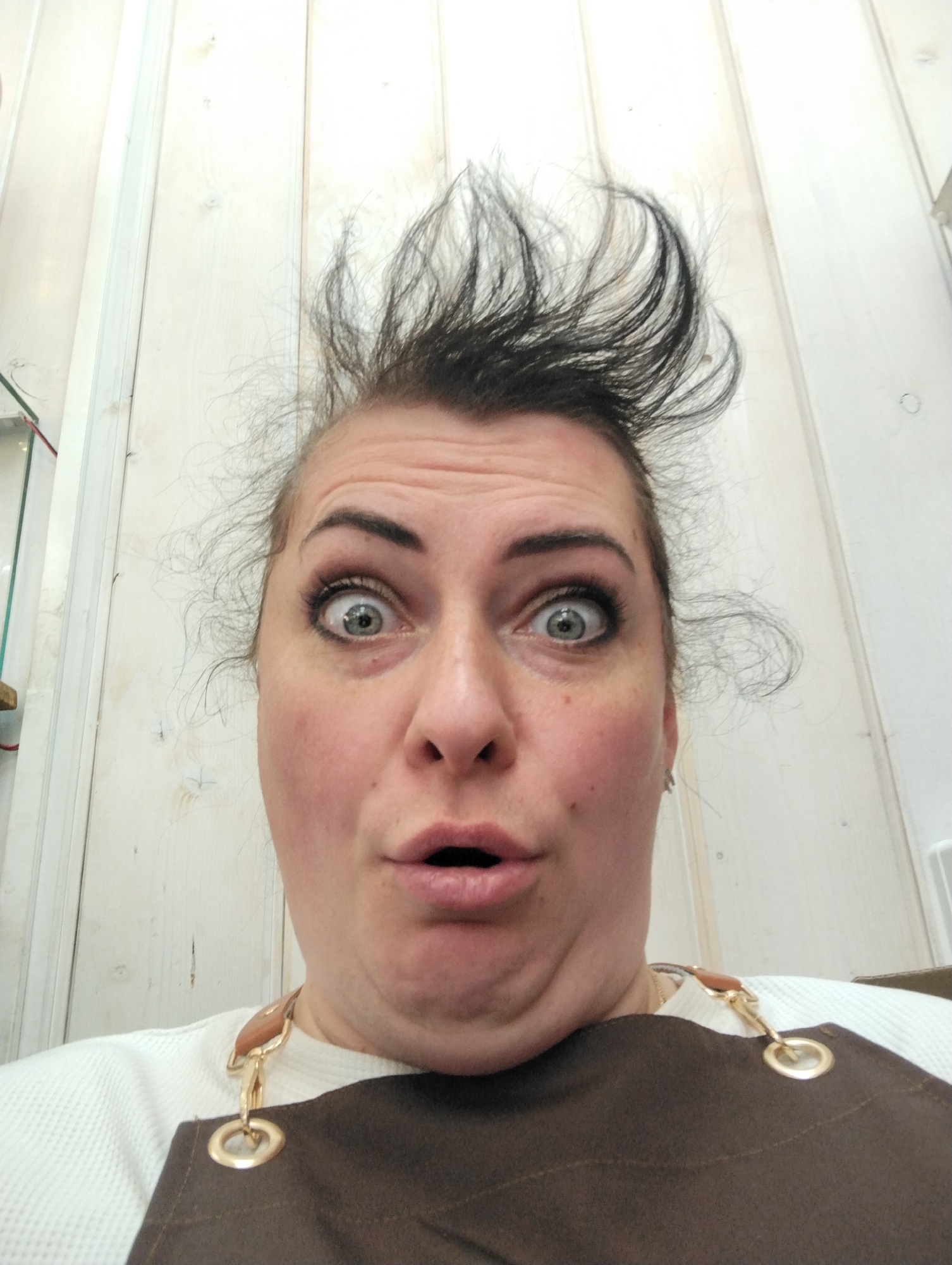 A woman is looking directly at the camera with a surprised expression. Her eyes are wide, and her mouth is open. Her hair is styled upwards in a messy fashion. She is wearing a white top and a brown apron with gold-colored rings and clips attaching the straps to the apron. The background consists of vertical, white wooden planks. There is no information in the image to determine a location in Votkinsk, Russia.