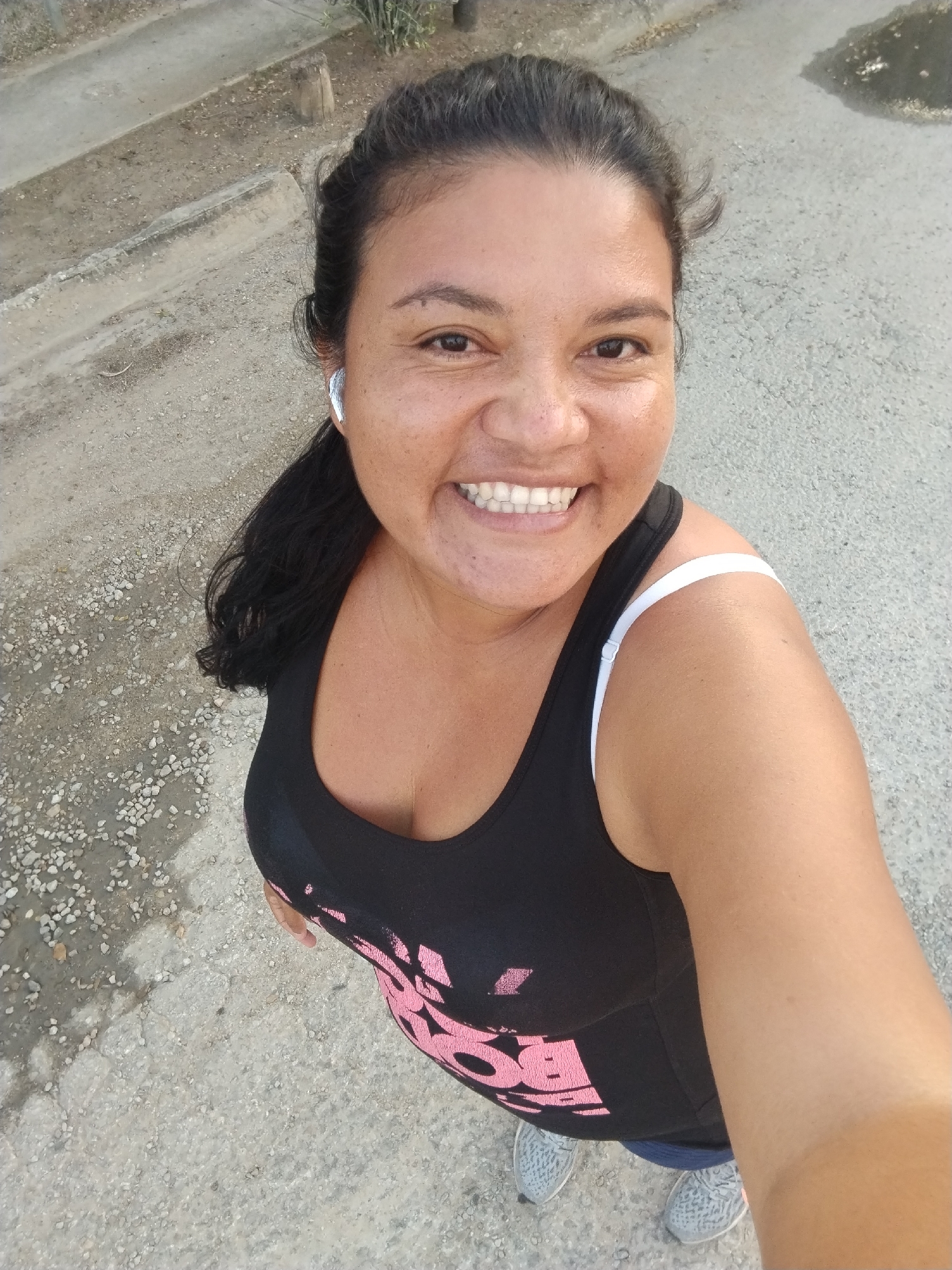 A medium-shot, high-angle, outdoor selfie shows a woman smiling at the camera. She is wearing a black tank top with pink lettering and grey athletic shoes. A white bra strap is visible. She has dark hair pulled back and is wearing a white earbud in her left ear. The ground is paved with asphalt and gravel. In the background, a concrete curb and some dry vegetation are visible. The scene is likely in Altagracia, Venezuela.