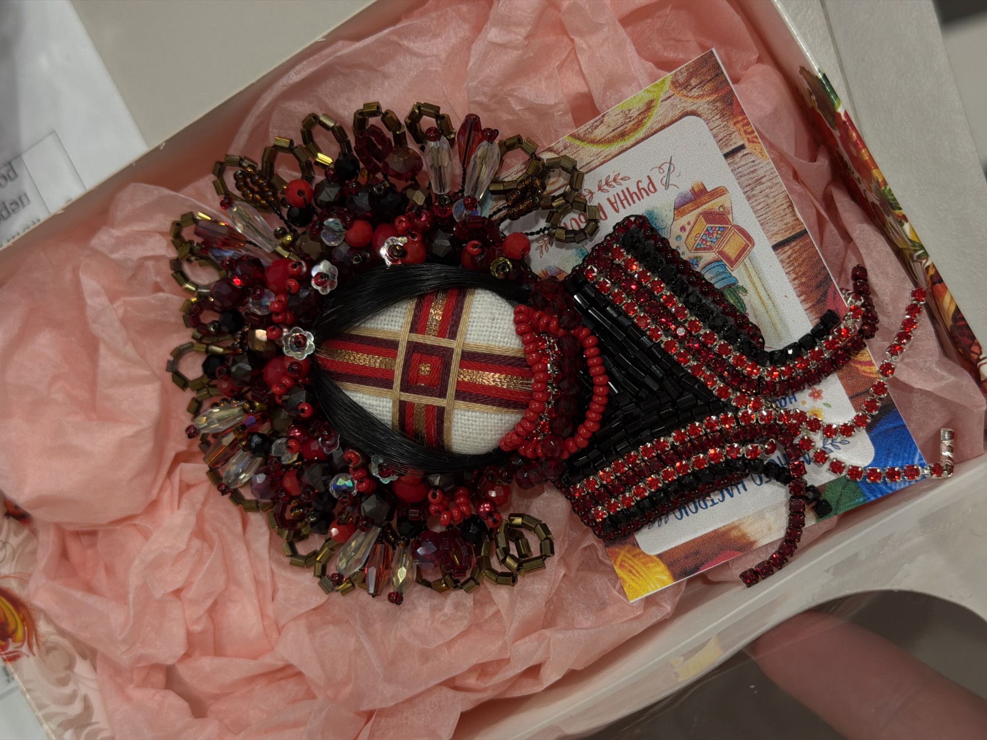 An overhead, medium shot captures a handcrafted, beaded brooch in the shape of a traditional Ukrainian headdress, known as a 'vinok' or 'maien'. The brooch is predominantly red, black, and gold, with intricate beadwork and bejeweled embellishments. It sits atop a pink tissue paper lining within a white box. The headdress features a central face-like element with a woven pattern, framed by a large, ornate circular arrangement of beads and crystals that mimics the floral and beaded decorations of a traditional headdress. Below the "face", a tiered, beaded collar and neckline are depicted, with dangling strands of red beads. The brooch is positioned on a small card with an illustration of a traditional Ukrainian folk art motif, featuring a stylized chest filled with colorful beads. The background is a soft pink, crinkled tissue paper, suggesting a gift presentation. The lighting is soft and diffused, highlighting the textures and colors of the brooch. The image's location is unknown, Unknown.