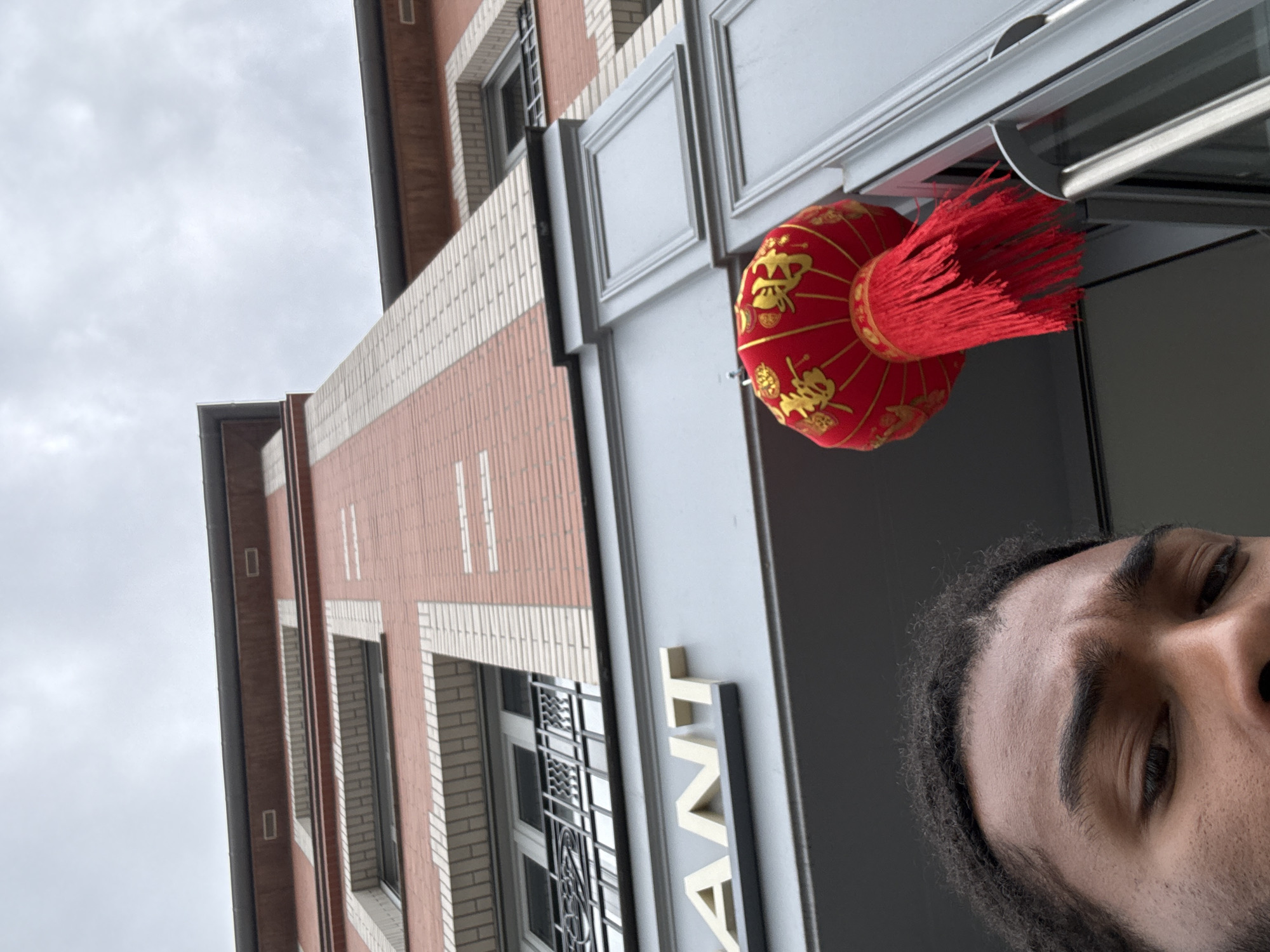 A low-angle outdoor shot in Domont, France shows a partial view of a man's face on the right, with black hair, dark eyes, and tan skin. A red Chinese lantern with gold details and a long red tassel hangs from the awning of a brick building. The awning is light gray with a sign that reads "ANT" in large, vertical gold letters. The brick building has white brick accents and several windows with black bars. The sky is overcast.