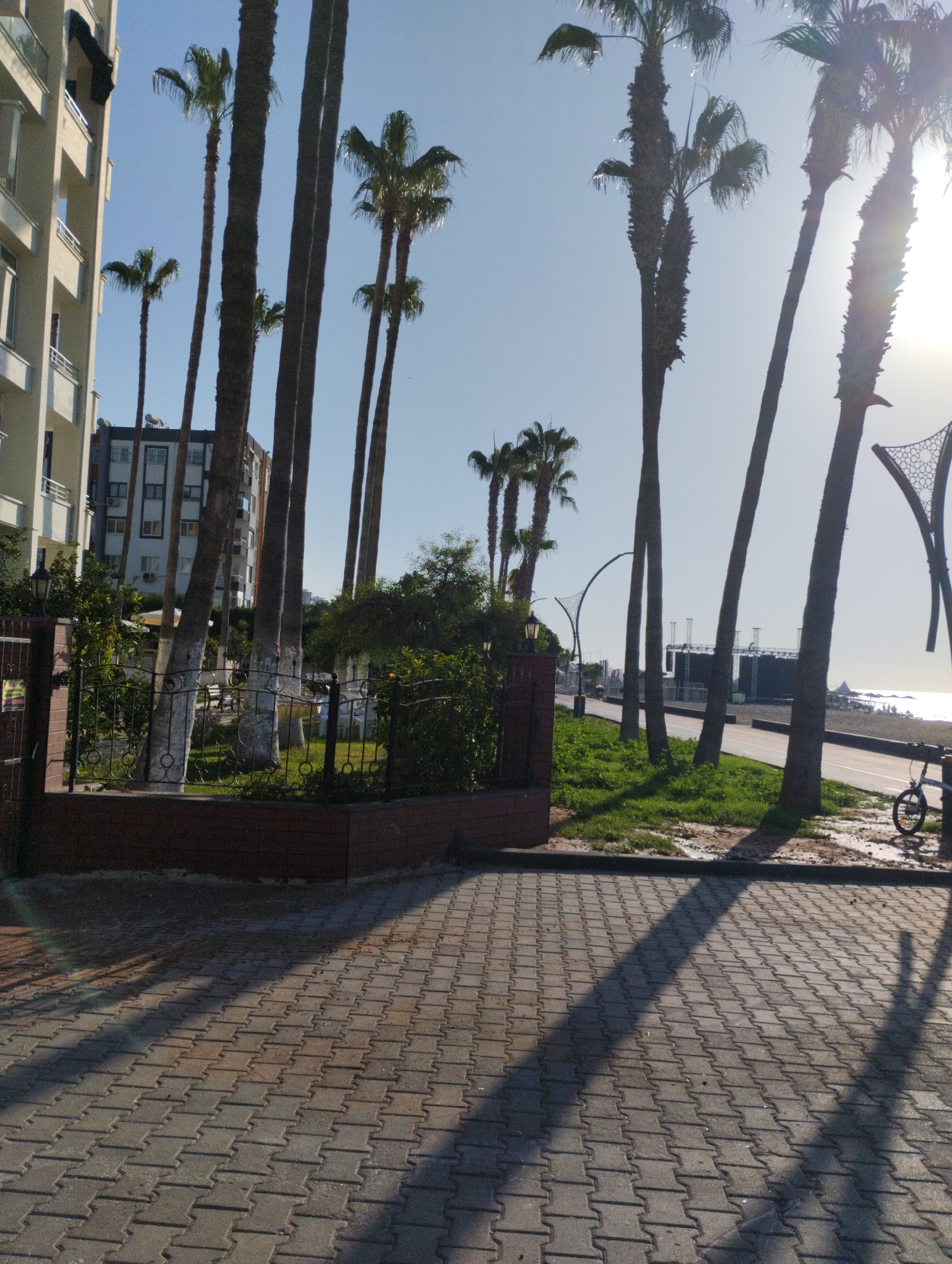The image depicts a street scene with palm trees, a building, and a beach in the background. The foreground is paved with interlocking concrete blocks, casting long shadows from the palm trees. A black metal fence runs along the side of the paved area, with a brick wall underneath. Beyond the fence, there is a grassy area with more palm trees and some greenery. In the background, a multi-story building with balconies is visible. Further in the distance, a beach with people and structures can be seen, bordering the sea. The sky is clear and bright, with the sun positioned high and slightly to the right, creating lens flare. The overall scene suggests a coastal urban environment, likely in a warm climate, consistent with the city of Mezitli, Turkey, known for its palm-lined streets and beaches.