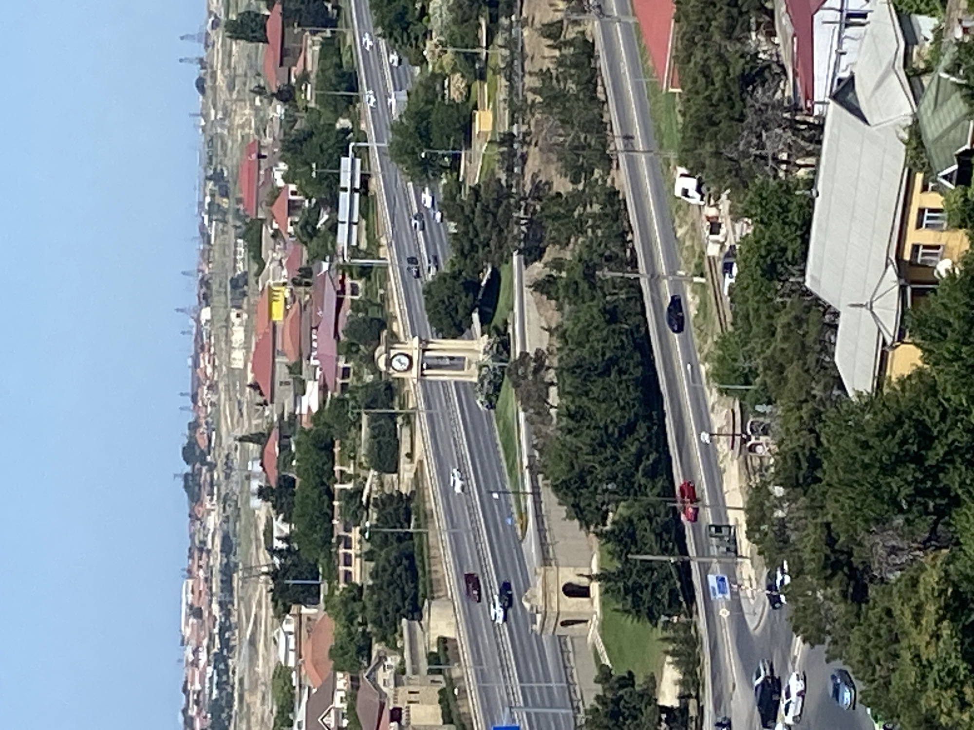 The image shows a high-angle view of a city street in Bakikhanov, Azerbaijan. A multi-lane highway runs through the center of the frame, with several cars and trucks visible on it. To the left of the highway, there is a clock tower and a small, ornate building with an arched entrance. Green trees and manicured lawns surround the highway and buildings. In the background, a cityscape with residential buildings and oil derricks can be seen under a clear blue sky. The overall impression is of a well-maintained urban environment with a mix of modern infrastructure and greenery.