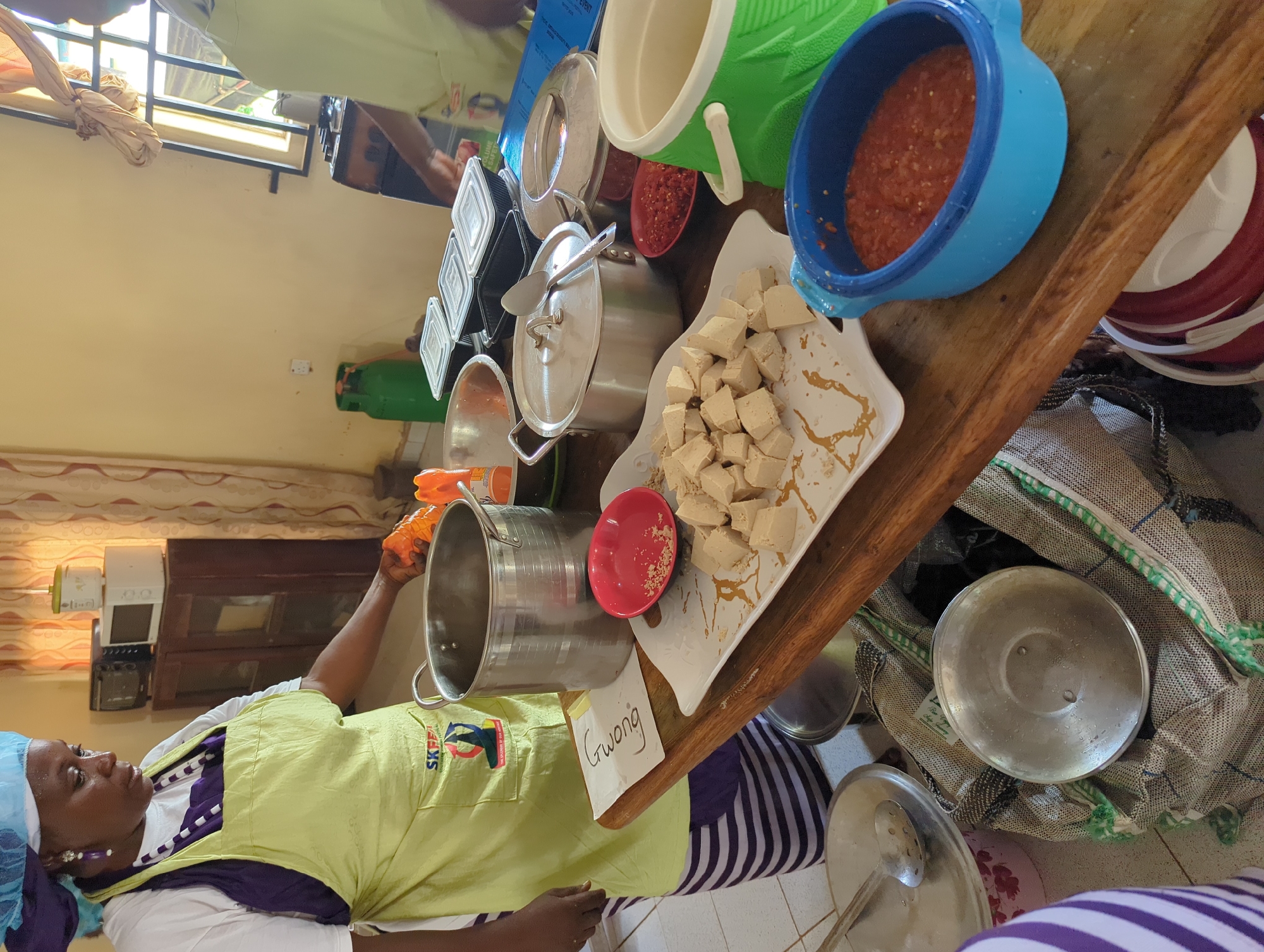 A woman in a light green apron and blue hair covering stands next to a wooden table inside a building in Maigizo, Nigeria. She's holding the lid of a stainless-steel pot. On the table, there's a plate of cut white cubes next to a small red bowl, various pots and containers. A piece of paper with "Gwong" written on it is also on the table. There's a gas cylinder near a stove along the wall in the background.