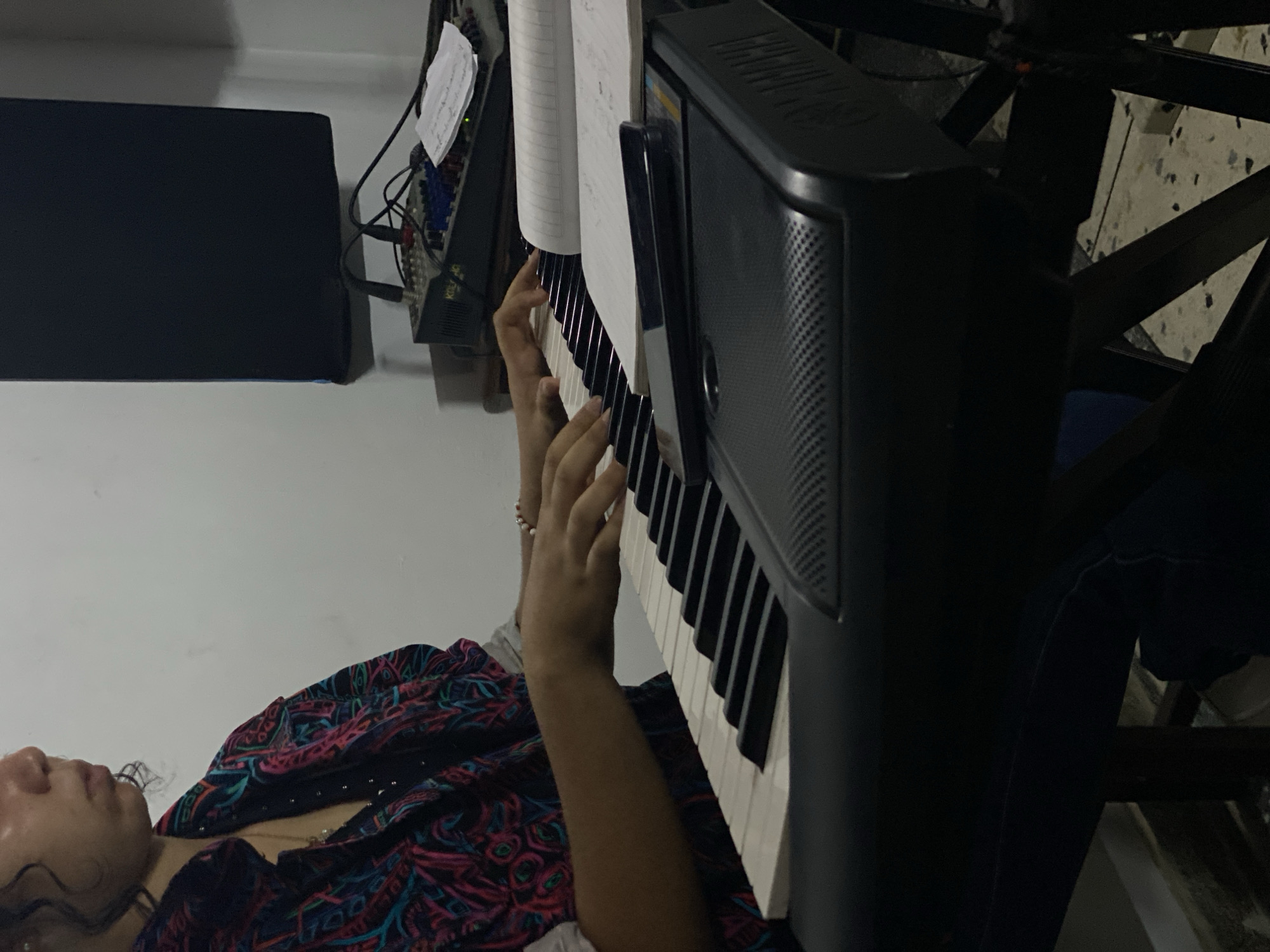 A person with fair skin and curly dark hair is lying down while playing a keyboard. The person is wearing a patterned top with dark and colorful designs. Their hands are positioned on the keyboard keys. Sheet music is visible above the keyboard. Adjacent to the keyboard, a mixing console or similar audio equipment is situated. A black monitor is to the left. A chair is visible on the right side of the keyboard. The exact location is Tinaquillo, Venezuela.
