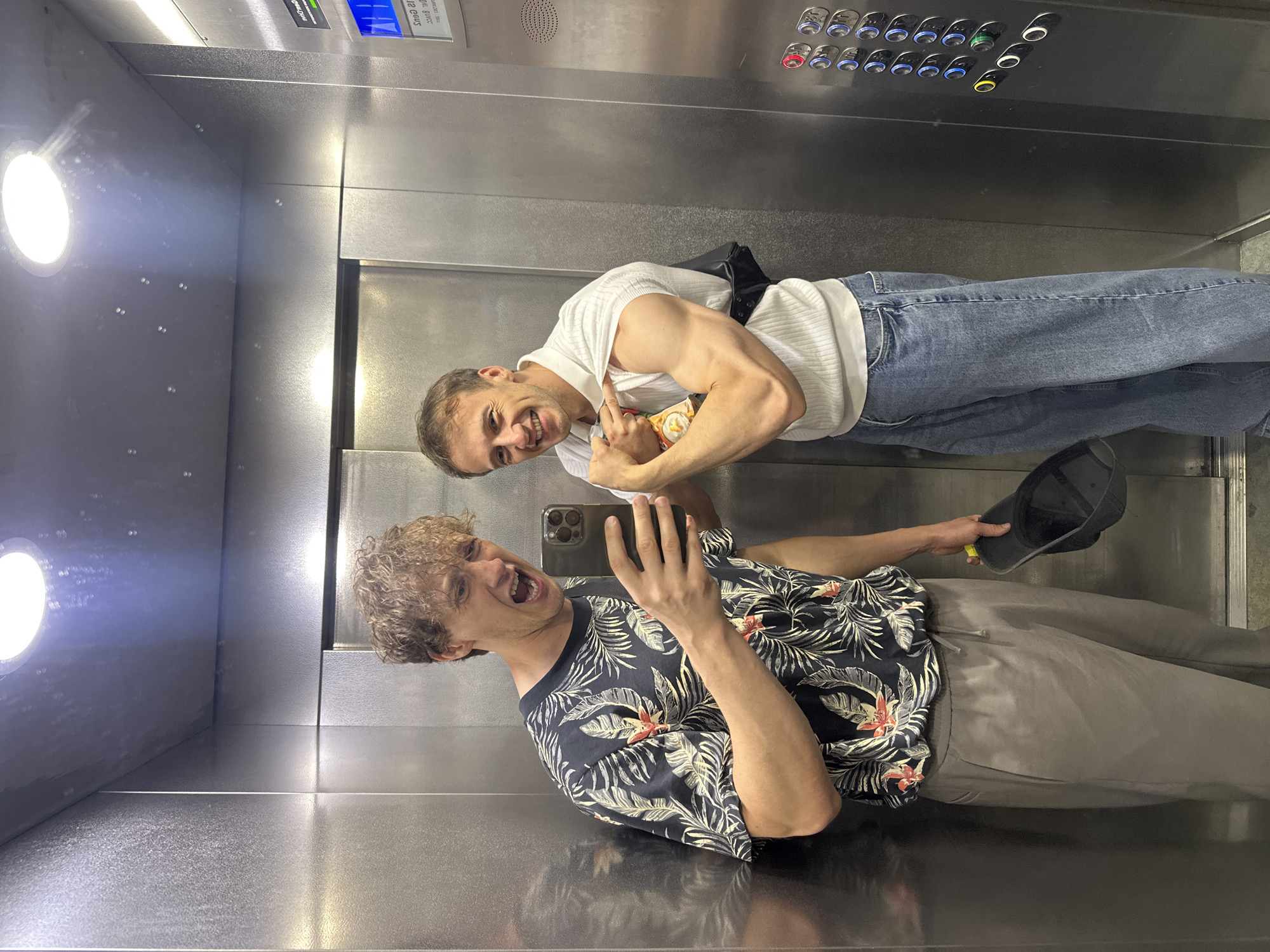In an elevator with stainless steel walls and a light panel on the ceiling, two young Caucasian men are taking a selfie. The man on the left, with curly blonde hair, is wearing a dark t-shirt with a floral pattern and grey pants, and is holding a smartphone in his right hand, taking the picture while shouting. The man on the right has short brown hair and is wearing a white short-sleeved polo shirt and blue jeans. He is flexing his right bicep, holding a snack bag in his left hand. An elevator control panel with illuminated buttons is visible on the right side of the image.