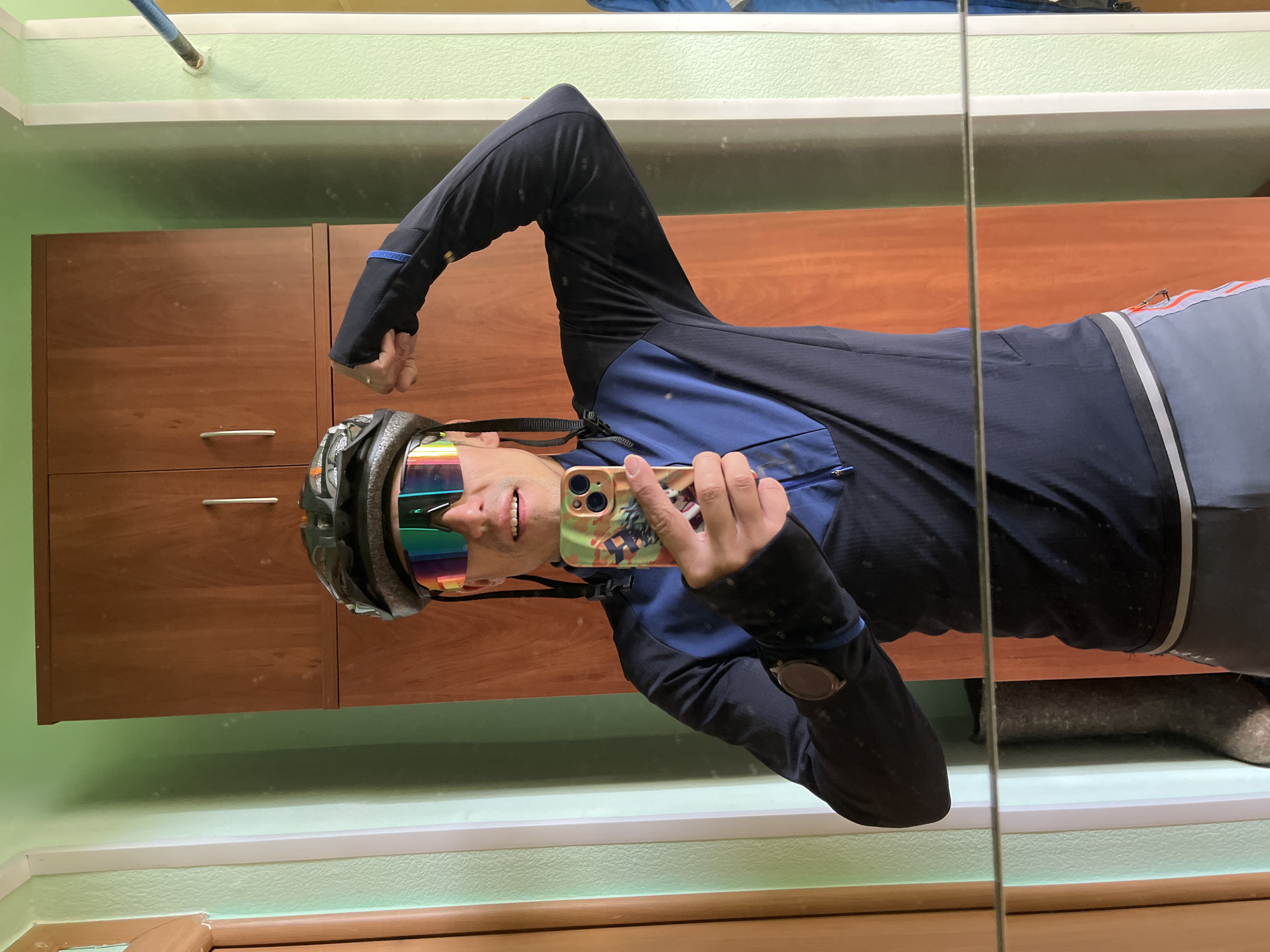 The image is a selfie of a person wearing a cycling helmet and iridescent sunglasses, taken in front of a mirror. The person is wearing a long-sleeved black and blue cycling jersey and grey cycling shorts. They are holding a smartphone with a patterned case. The background includes a wooden cabinet and a light green wall with white trim. The reflection suggests the photo was taken indoors. It is not possible to determine the specific location in Chaikovsky, Russia from the given image.