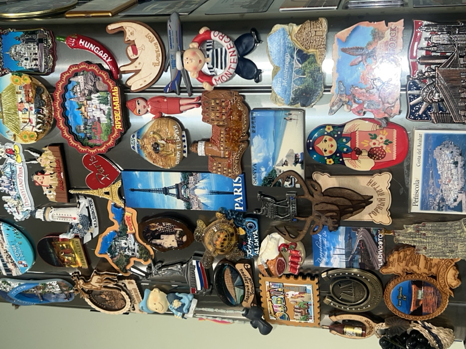 The image shows a collection of travel magnets attached to a metallic surface. Destinations include Hungary, Paris, Sanya, Montenegro, Egypt (Sharm El Sheikh), Yaroslavl, Disneyland Paris, Jerusalem, Sochi, and Volgograd. There are magnets of cityscapes, cultural icons like matryoshka dolls, and regional symbols. Specific locations within these cities, such as Peñíscola (Costa del Azahar) and Gorny Altai, are also represented. Some magnets have place names written in Cyrillic script.
