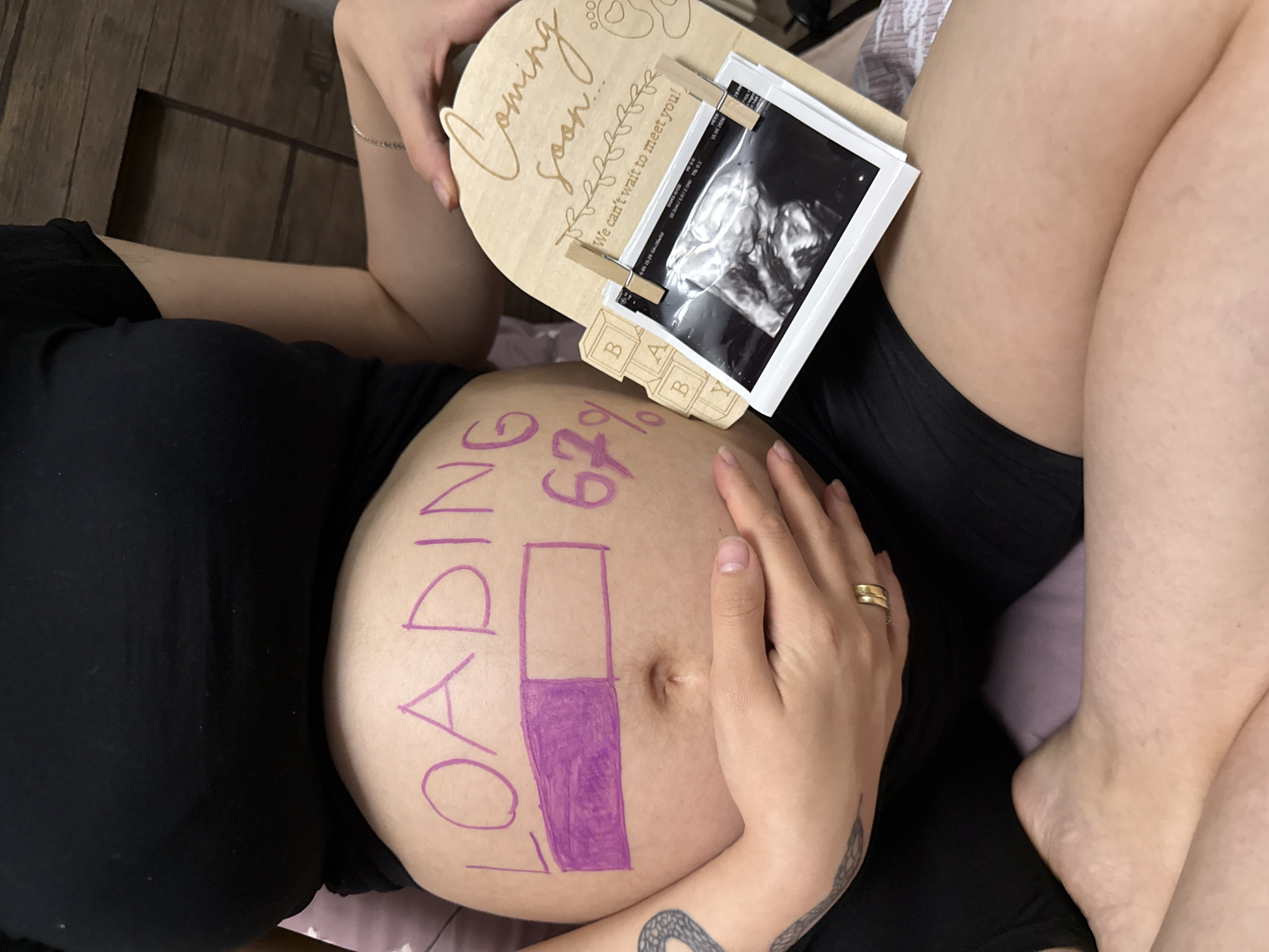 In a dimly lit indoor setting, a pregnant person is seated, cradling their baby bump which is adorned with the text "LOADING 67%" written in purple marker, accompanied by a progress bar indicating completion. To the right, a wooden plaque with "Coming soon! We can't wait to meet you!" is propped up, holding an ultrasound image and wooden blocks spelling "BABY." The scene appears to be a personal announcement of a pregnancy. There is no information in the image to indicate a location in Tbilisi, Georgia.