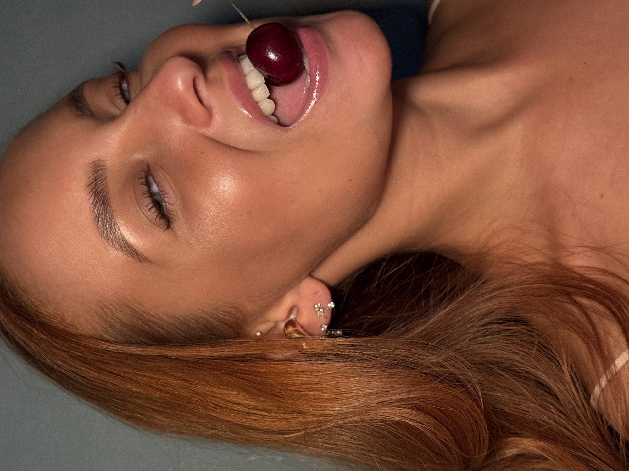 The image shows a close-up, waist-up shot of a young woman with auburn hair, holding a cherry in her mouth. She is wearing subtle jewelry on her ear and a white garment, possibly a top. Her skin appears to have a dewy, sun-kissed glow. The background is a plain, muted blue-gray color. The lighting suggests an indoor setting, possibly with artificial light highlighting her features. There is no indication of the city of Chaikovsky, Russia, or any other specific location.