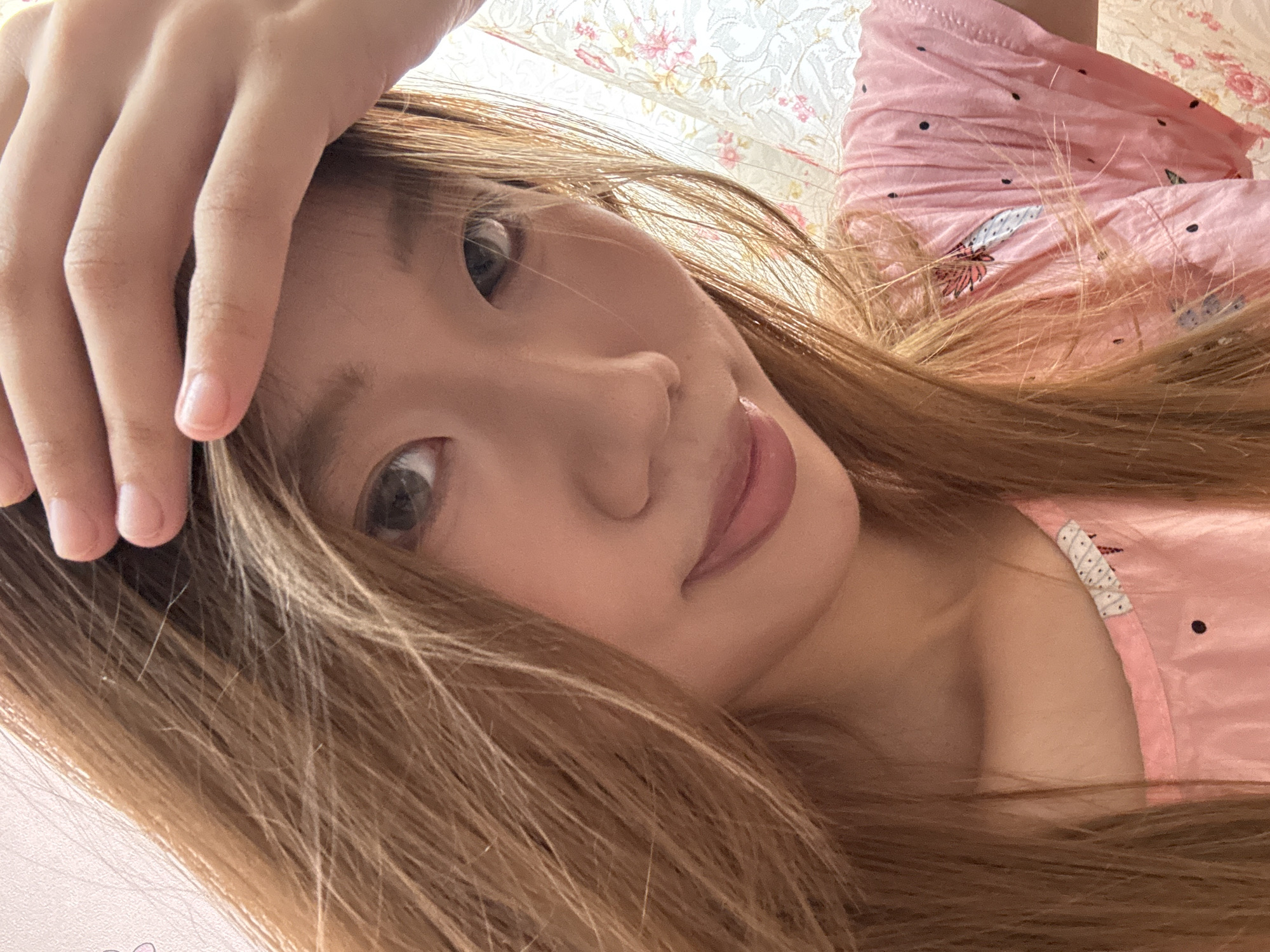A close-up, selfie-style photograph shows a young East Asian woman with long, blonde-brown hair pushed back from her forehead by her right hand. She is wearing a pink t-shirt with a floral pattern. The background appears to be an indoor setting with a textured wall to the left and a floral curtain to the right. The woman is looking at the camera with a slight smile. The lighting suggests the photo was taken during the daytime.