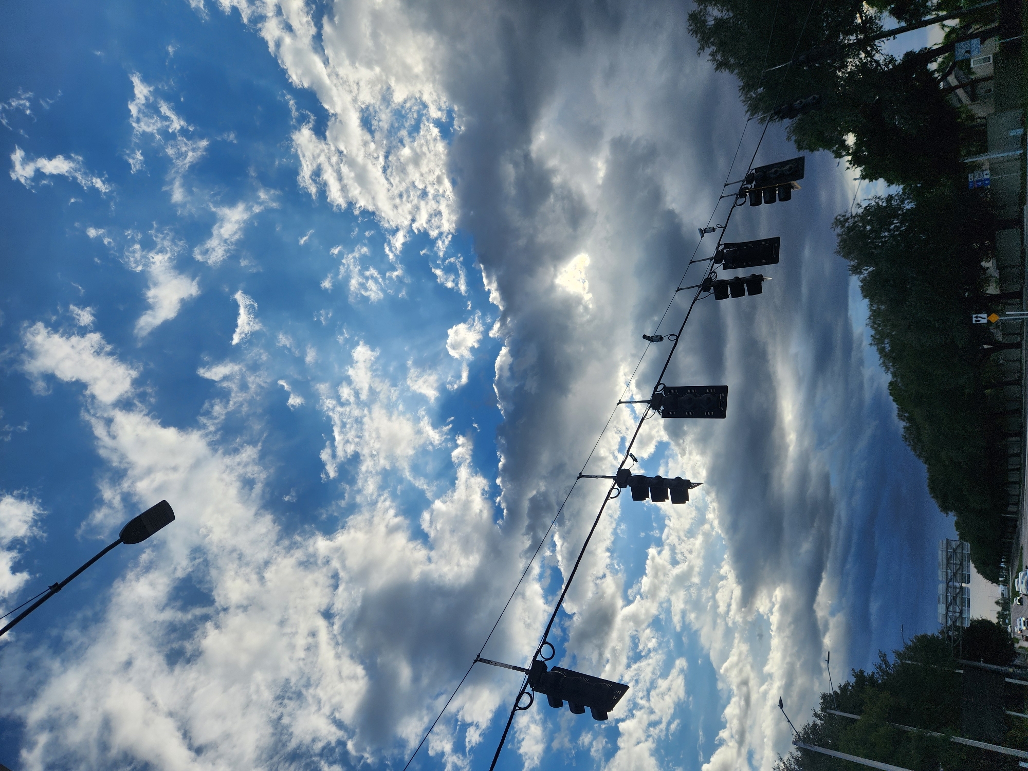 The image shows a street scene in Maitland, Florida, USA, with a dramatic sky filled with both bright white and dark grey clouds. Overhead, a series of traffic lights hang from wires across the road, silhouetted against the sky. Some of the traffic lights appear dark, while others show signs of illumination. Trees line both sides of the road, and a few vehicles are visible on the street below. The overall impression is one of an approaching storm or the aftermath of one, with the play of light and shadow adding to the atmospheric effect.