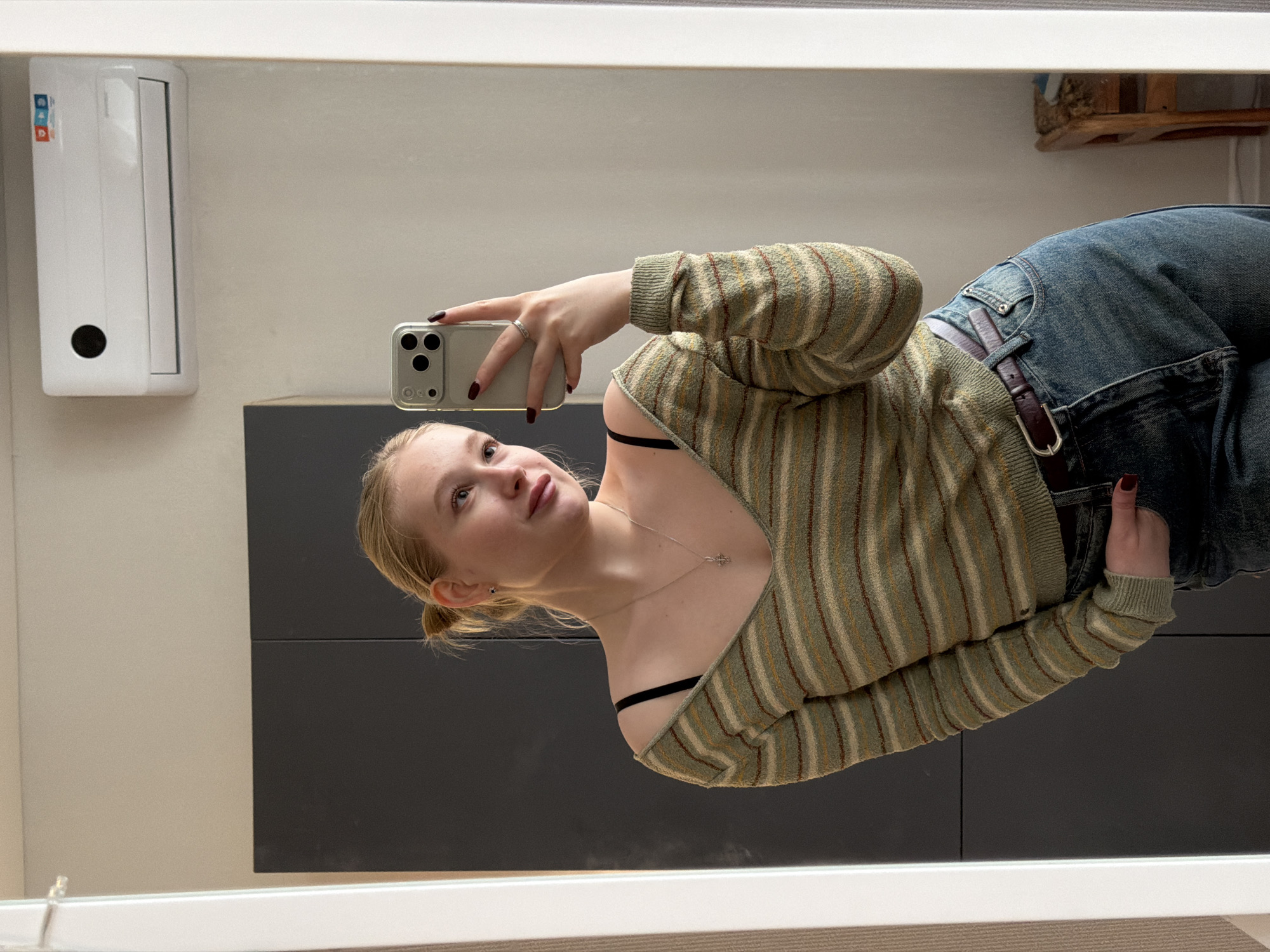 A young woman is taking a selfie in a mirror. She is wearing a striped sweater and blue jeans with a brown belt. She is holding a smartphone in her right hand, and her left hand is on her hip. She has blonde hair pulled back in a ponytail and is wearing a necklace. The background includes a white wall, a black cabinet, and an air conditioner unit. The location is possibly in Засечное, Russia.