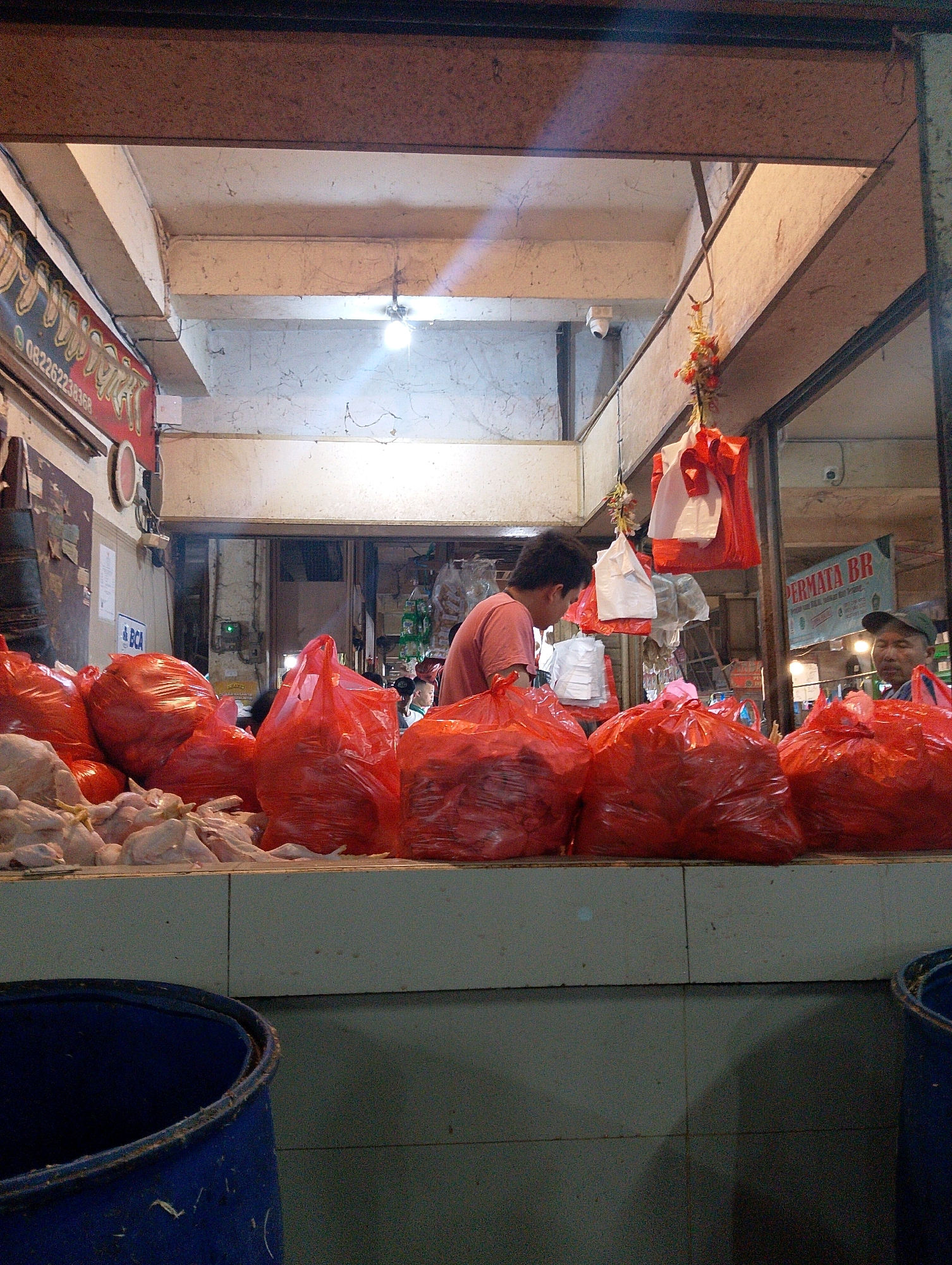 The image depicts a market scene in Lembang, Indonesia. In the foreground, a row of uncooked chickens is displayed on a counter, some partially covered with plastic. Behind them, large red plastic bags filled with unknown contents are piled up. To the left, a blue plastic bin is visible. In the middle ground, a man wearing a pink t-shirt is standing, looking down towards the counter. Above him, red and white plastic bags are hanging, along with some decorative elements. To the right, another man wearing a green cap is visible, partially obscured by a red plastic bag. The background shows a market stall with various items and signage, including a red sign with text and a green banner that reads "PERMATA BR". The environment appears to be an indoor market with exposed ceiling beams and lighting fixtures.