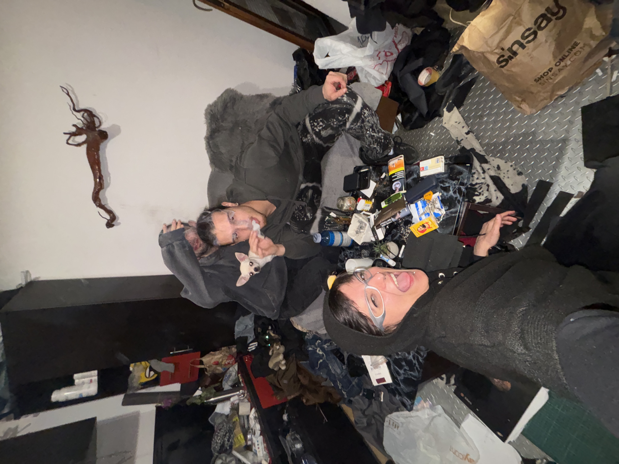 Two people are lying on a cluttered floor in what appears to be an interior space. One person is holding a small dog. The floor is covered with clothes, packaging, and other miscellaneous items. In the background, there are bags and objects suggesting a cluttered living space. The scene contains no immediate clues to Sofia, Bulgaria.