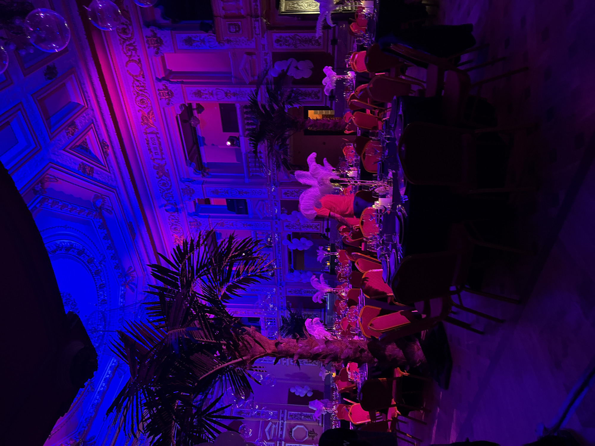 This image captures a lavish indoor event space, likely a grand ballroom or hall, illuminated by vibrant blue and magenta uplighting. The perspective is tilted, showing the ornate ceiling and walls adorned with intricate gold moldings and decorative panels.
The room is set up with multiple round tables, each draped in dark tablecloths and surrounded by chairs with distinctive red or orange upholstered backs and seats. Each table appears to be set for guests, featuring an array of glassware, including wine glasses and tumblers. White, feather-like decorations, possibly centerpieces, are visible on some tables, adding to the festive atmosphere.
Several artificial palm trees are strategically placed throughout the space, contributing to a tropical or exotic theme amidst the opulent architecture. Transparent, spherical decorations, resembling bubbles or unique light fixtures, hang from the high ceiling, catching the colorful glow.
The scene suggests an evening event, such as a gala, reception, or private party, given the elaborate setup and dramatic artificial lighting. No people are clearly visible, indicating the photograph might have been taken before guests arrived or after they departed.
Notable architectural details include grand arches and meticulously decorated ceilings and upper walls, characteristic of historic European buildings. One cropped image reveals the text "15-11-1913" inscribed on a wall, likely a significant date related to the venue's history or a past event. The overall mood conveyed is one of luxury, festivity, and grandeur. The location context provided is Sofia, Bulgaria.