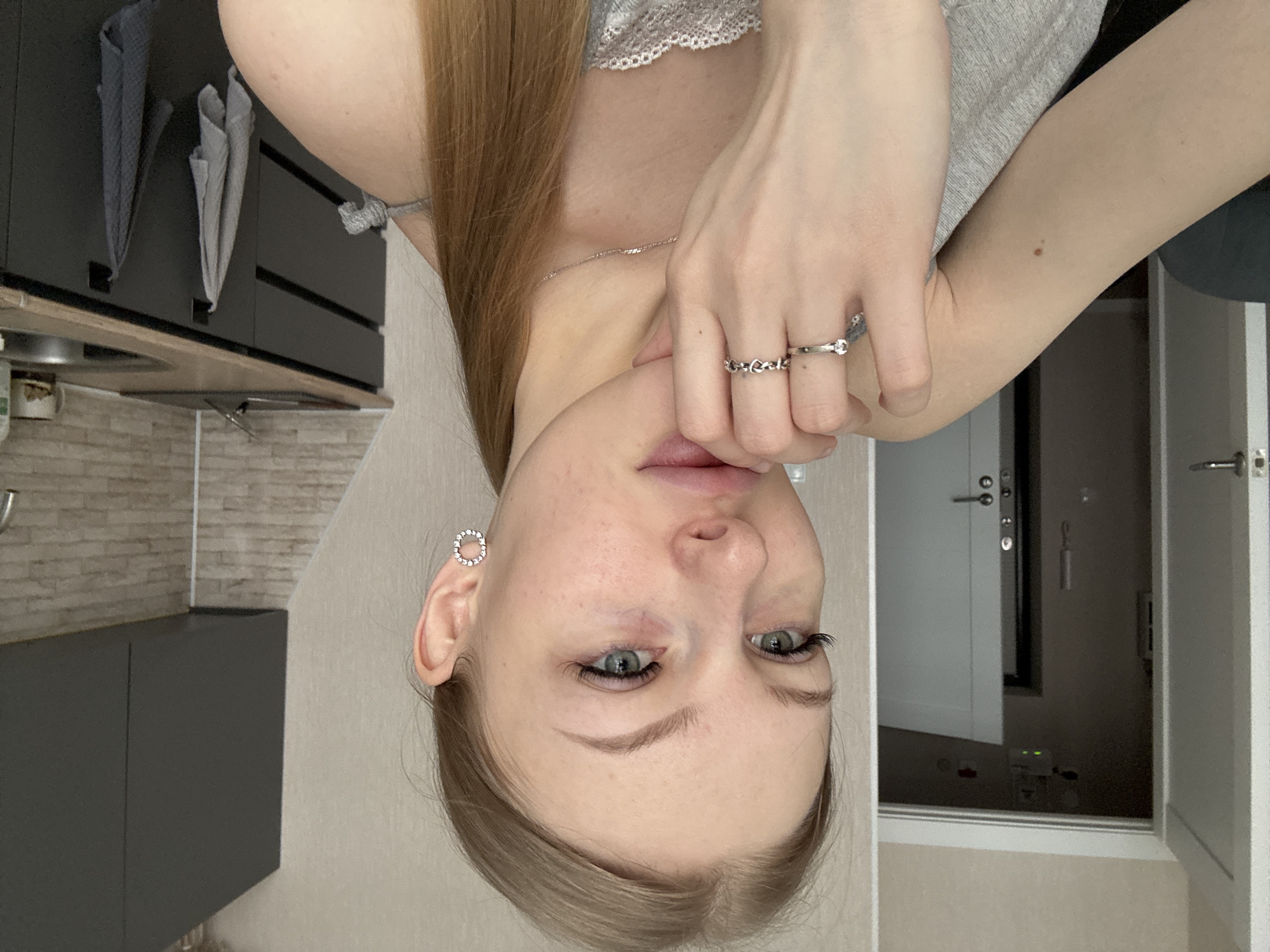 A young adult female is depicted in a close-up indoor photograph, primarily from the chest up. She has light brown hair, pulled back into a ponytail visible over her right shoulder, and light-colored eyes. Her complexion is fair. She wears a light grey camisole with lace detailing on the neckline. Her jewelry consists of silver-toned hoop earrings adorned with small clear stones, a thin silver-toned chain necklace, and two silver-toned rings on her left ring finger: one with a single clear stone and another with a repeating heart pattern. Her right hand is raised, with fingers positioned near her lips. A small, dark brown nevus is visible on her upper right arm.
The background setting is an interior residential space. To the subject's right, a modern kitchen area features dark grey flat-panel lower and upper cabinets with black bar handles, a light wood-toned countertop, a stainless steel sink with a chrome faucet, and a light-colored rectangular brick-patterned backsplash. Two textile kitchen towels, one light grey and one dark grey, hang from hooks on the lower cabinets. To the subject's left, a partially open white door with a silver-toned handle provides a view into a hallway. In the distant hallway, another white door is partially visible, along with a wall-mounted intercom system and a fire alarm/smoke detector exhibiting a green indicator light on the ceiling. The surrounding walls are light beige. The precise location within the city of Perm, Russia cannot be determined from the visual information present in the image.