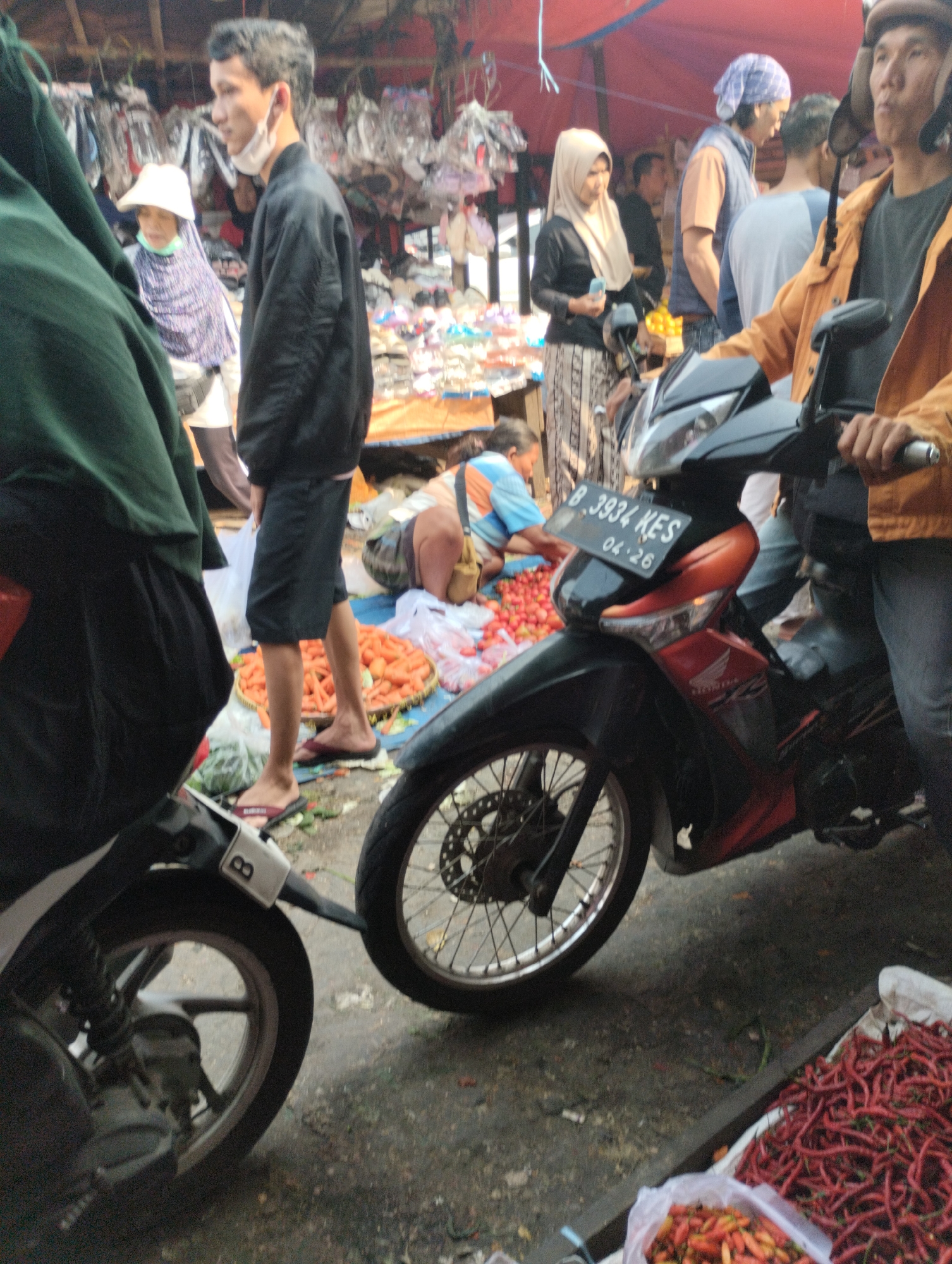 This is a medium shot, eye-level, horizontal photograph taken in a market setting in Bekasi, Indonesia. The foreground features the front of a black motorcycle with red accents. To the left, a person wearing a green garment is partially visible, obscuring the motorcycle. In the midground, several people are engaged in market activities. A man in a black jacket and shorts is standing, facing right, with a white face mask around his neck. To his left, a woman in a white headscarf and a purple patterned garment is also standing. Further into the midground, a person in a blue and orange shirt is sitting down, surrounded by baskets of produce, including carrots and tomatoes. In the background, market stalls are visible with various goods hanging and displayed. The ground is a textured, wet asphalt or concrete. The scene appears to be outdoors, under a covered market area as suggested by the presence of red tarpaulin in the upper background. The overall lighting suggests it is daytime.