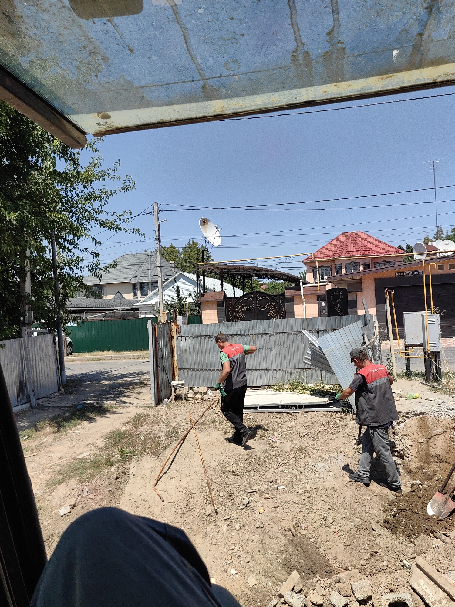 The image depicts an outdoor scene in Shymkent, Kazakhstan, on a sunny day. In the foreground, a person's leg, clad in dark pants, is visible from a low vantage point, suggesting the viewer is seated. The ground is a mix of dirt, small stones, and some debris.
In the middle ground, two individuals wearing work vests over dark shirts and pants are engaged in manual labor. One person, on the left, is holding what appears to be a rake or broom and moving away from the viewer. The other individual, on the right, is in the process of lifting and carrying a bent piece of corrugated metal siding, which is partially damaged. Both individuals are wearing gloves, indicating a work environment.
The background reveals a residential area with houses of varying architectural styles and roof colors, including a prominent house with a red-tiled roof. Fences, including a green metal fence and a corrugated metal fence, separate properties. Power lines and a satellite dish are visible against a clear blue sky. A street is visible in the left background, with a parked white vehicle. A gas meter and yellow piping are also present on the right side of the scene, attached to a building. The overall environment appears to be a construction or repair site in a suburban or semi-urban setting within the city.