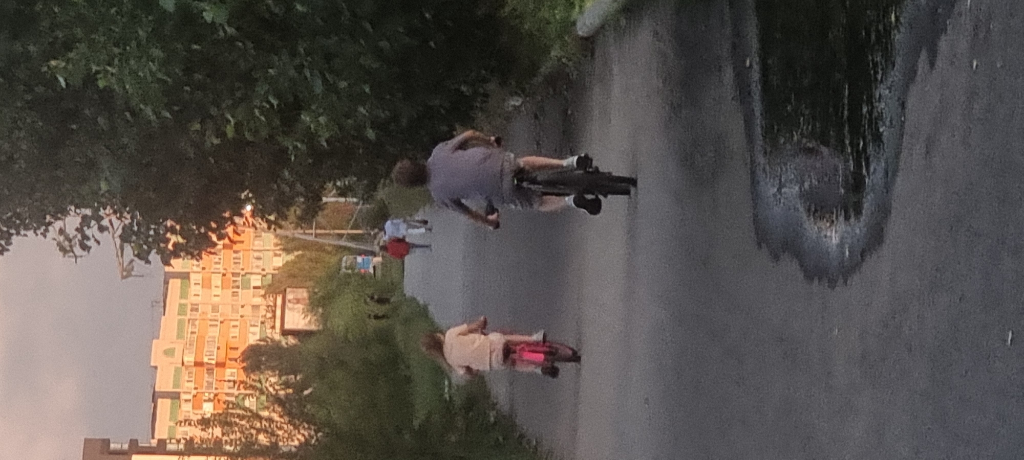 The image depicts a paved pathway in Aktobe, Kazakhstan, likely a park or residential area, at dusk or dawn, given the soft lighting. Two children are riding bicycles on the path. In the foreground, a child with long hair, wearing a light-colored shirt and shorts, is on a pink bicycle, facing away from the camera. To the right and slightly ahead, another child, also facing away, is riding a bicycle. This child wears a gray t-shirt and shorts.
In the middle ground, several people are walking away from the camera on the same path. One individual in a red jacket is particularly noticeable, accompanied by another person in a light-colored top. Further in the distance, behind a sign and trees, a multi-story building complex with orange and beige facades is visible, characteristic of Soviet-era architecture found in cities like Aktobe. The path is bordered by dense green foliage and trees on the right side of the image, with more trees and the building complex on the left. A puddle of water is visible on the pathway in the lower right portion of the image, reflecting some light. A utility pole with wires is also present on the left side of the path.