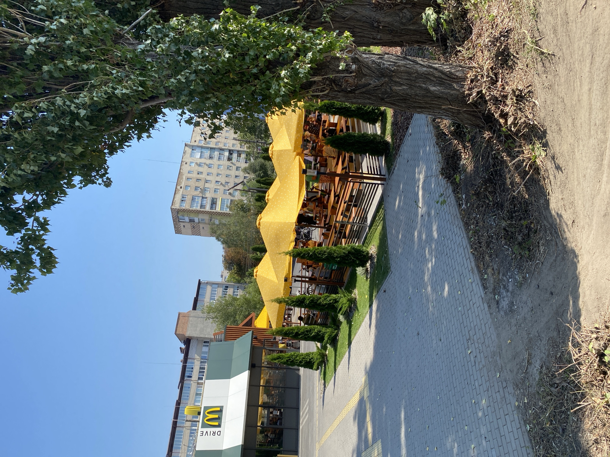 The image shows a McDonald's restaurant with a drive-thru lane in Vinnytsia, Ukraine. Outside the restaurant, there is a seating area with yellow umbrellas and wooden tables and benches. Several people are seated at the tables, and some are walking in the background. The restaurant building is visible, with the McDonald's logo prominently displayed. The area is paved with gray bricks, and there are trees and green grass surrounding the seating area. In the background, there are multi-story apartment buildings. The sky is clear and blue.
