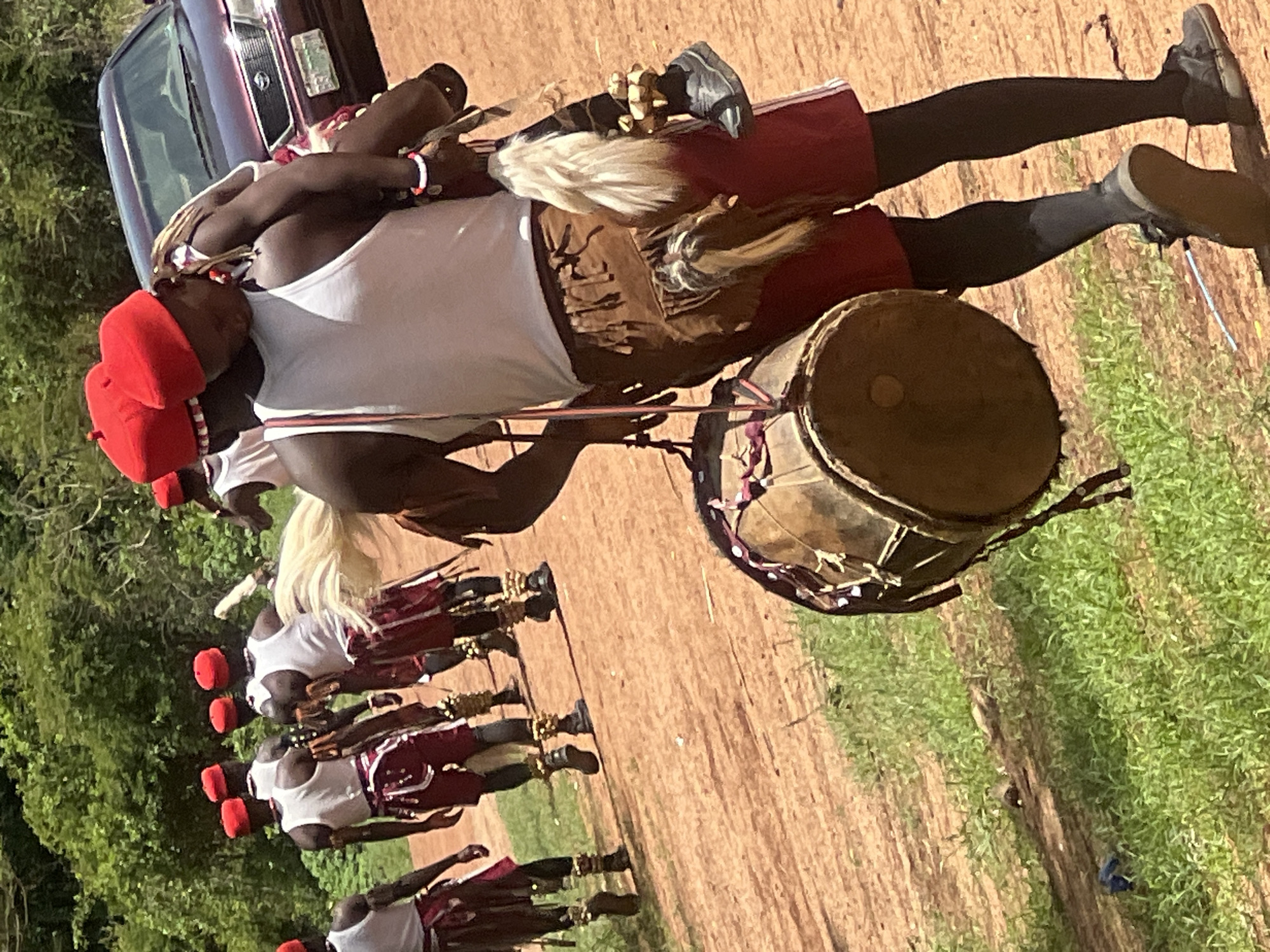 The image shows a procession of men in Jos, Nigeria, seemingly captured outdoors on a dirt path with green vegetation to the side. The men are dressed in red hats, white tank tops, and red shorts with brown fabric accents, and anklets. One man carries a large drum suspended by a pole resting on his shoulders. Another man is visible near a burgundy car, possibly accompanying the group. The image appears to be taken from a slightly elevated perspective.
