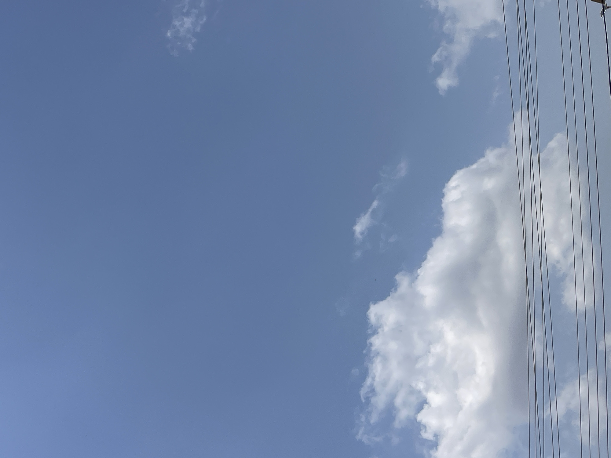 The image depicts a clear blue sky with scattered white clouds, as viewed from Rafin Par, Nigeria. In the foreground, several dark power lines are stretched horizontally across the frame. The scene is devoid of any people or specific structures, focusing solely on the natural elements of the sky and the man-made power lines.