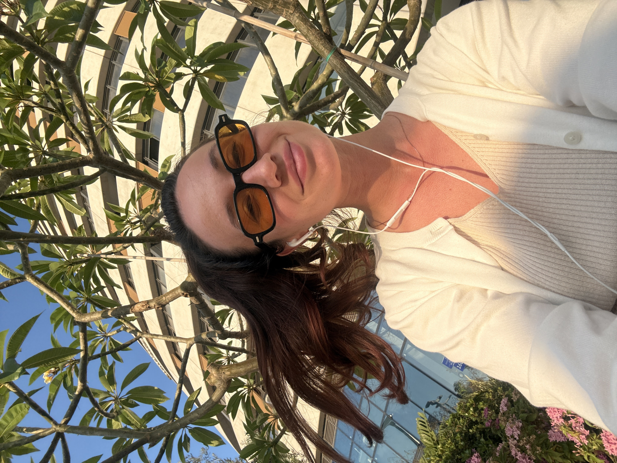 The image is a selfie taken outdoors in Tel-Aviv, Israel. The subject is a woman with dark hair tied in a ponytail, wearing orange-tinted sunglasses, a white cardigan over a ribbed cream top, and white earbuds. She is smiling slightly. Behind her, there is a tree with green leaves and branches, and a modern building with horizontal bands of windows and beige siding. The sky is clear blue. In the lower left of the image, there is a bush with small pink flowers. The lighting suggests it is late afternoon. The building appears to be located in a commercial or residential area of the city.