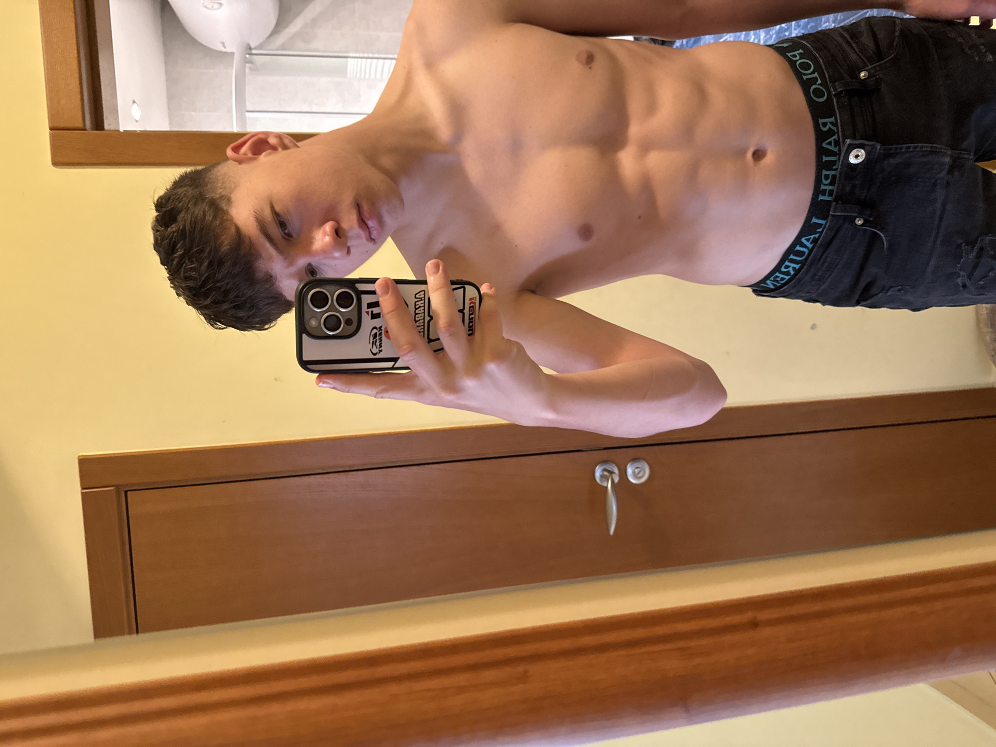 A shirtless young man with short dark hair takes a selfie in front of a mirror. He is wearing dark jeans with a visible "RALPH LAUREN" waistband. His face is partially visible as he holds a phone with a patterned case. The background shows a yellow wall, a brown wooden door with a handle, and a glimpse of a bathroom with a white toilet. The image does not provide sufficient information to determine the location within Gabrovo, Bulgaria.