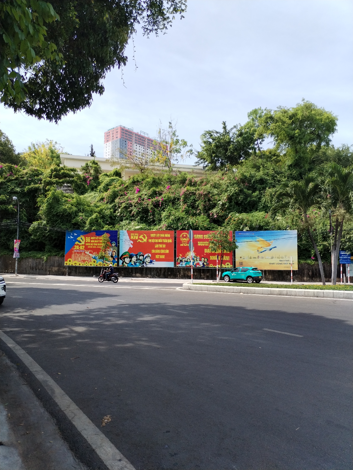 The image captures a street scene in Bac Nha Trang Ward, Vietnam. A wide asphalt road occupies the foreground, with shadows from overhead trees dappling its surface. A white line marks the edge of the road on the left. To the right of the road, a paved area with a low white curb separates it from a retaining wall.
Along the retaining wall, a series of large billboards are displayed. The leftmost billboard features a red background with yellow accents, depicting sunflowers and a group of people in traditional attire. The central billboard also has a red background with yellow elements, showing more figures in uniform. The rightmost billboard is predominantly blue with images of airplanes and clouds, alongside text.
Behind the billboards, a steep, green, and overgrown hillside is visible, dotted with pink flowers. A tall, multi-story building with a pink and gray facade is perched atop the hill. Trees with dense green foliage surround the building and the hillside.
In the street, a dark-colored motorcycle with a rider is in motion. Further down the road, a teal-colored car is parked. A street lamp stands on the left side of the road, and a street sign is visible on the right. The sky above is a pale blue, with a few wispy clouds.