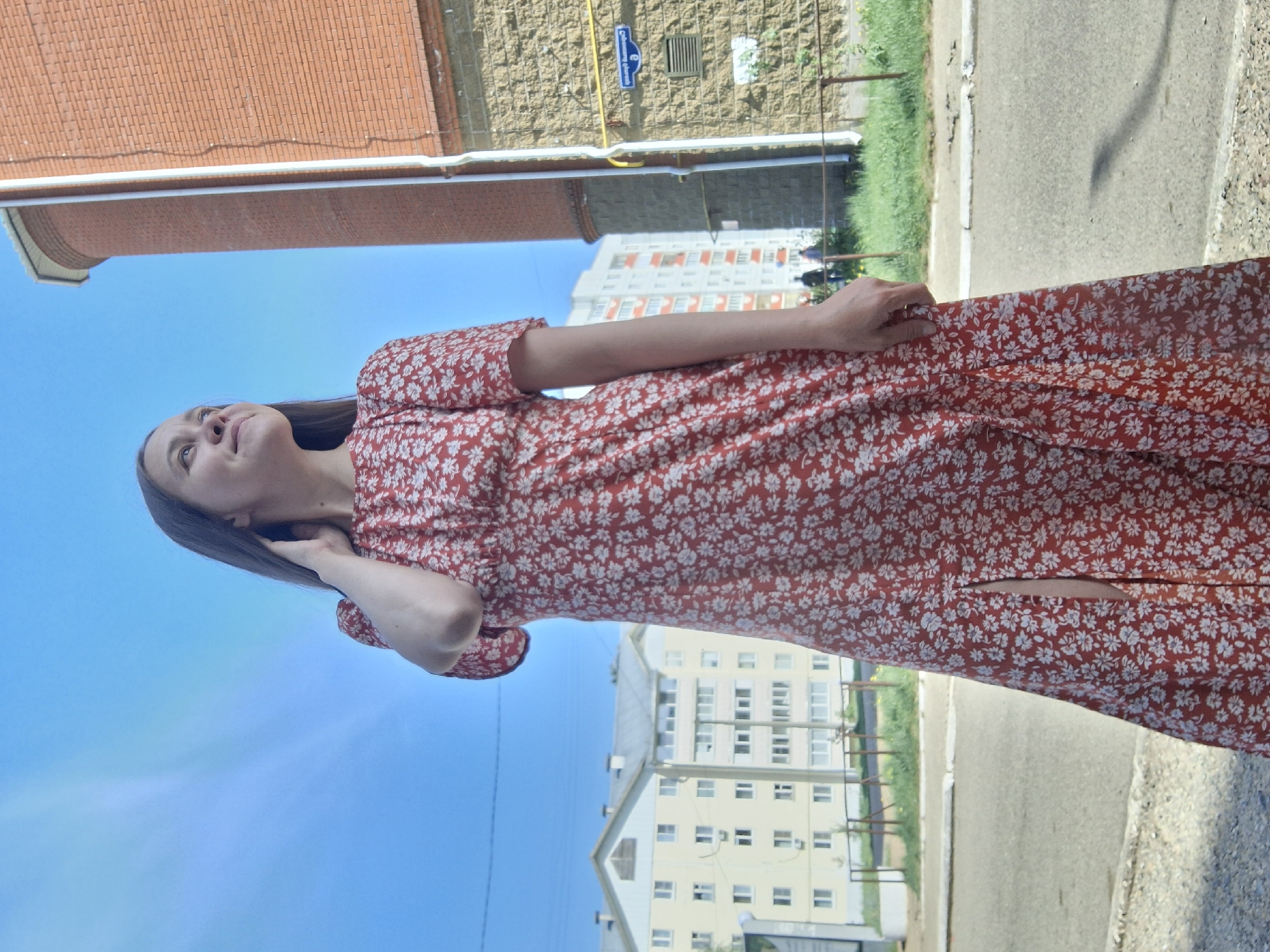 A woman is standing on a city street in Chaikovsky, Russia. She is wearing a red, floral-patterned dress with a slit. The sky is blue and clear. Buildings surround her, including brick buildings and a taller white building. There is a street sign visible on one of the buildings. There is also a sidewalk, street, and some greenery.