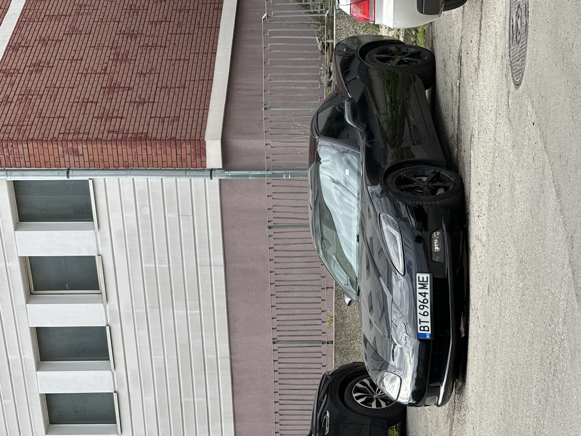 A black Chevrolet Corvette is parked on a paved surface in front of a building in Veliko Tarnovo, Bulgaria. The building has a facade of white horizontal siding and a section of red brick. A metal fence runs along the front of the building. The Corvette has a license plate that reads "BT 6964 ME". Another car is partially visible on the left, and a white car is visible on the right. The sky is overcast and reflected in the car's windshield.