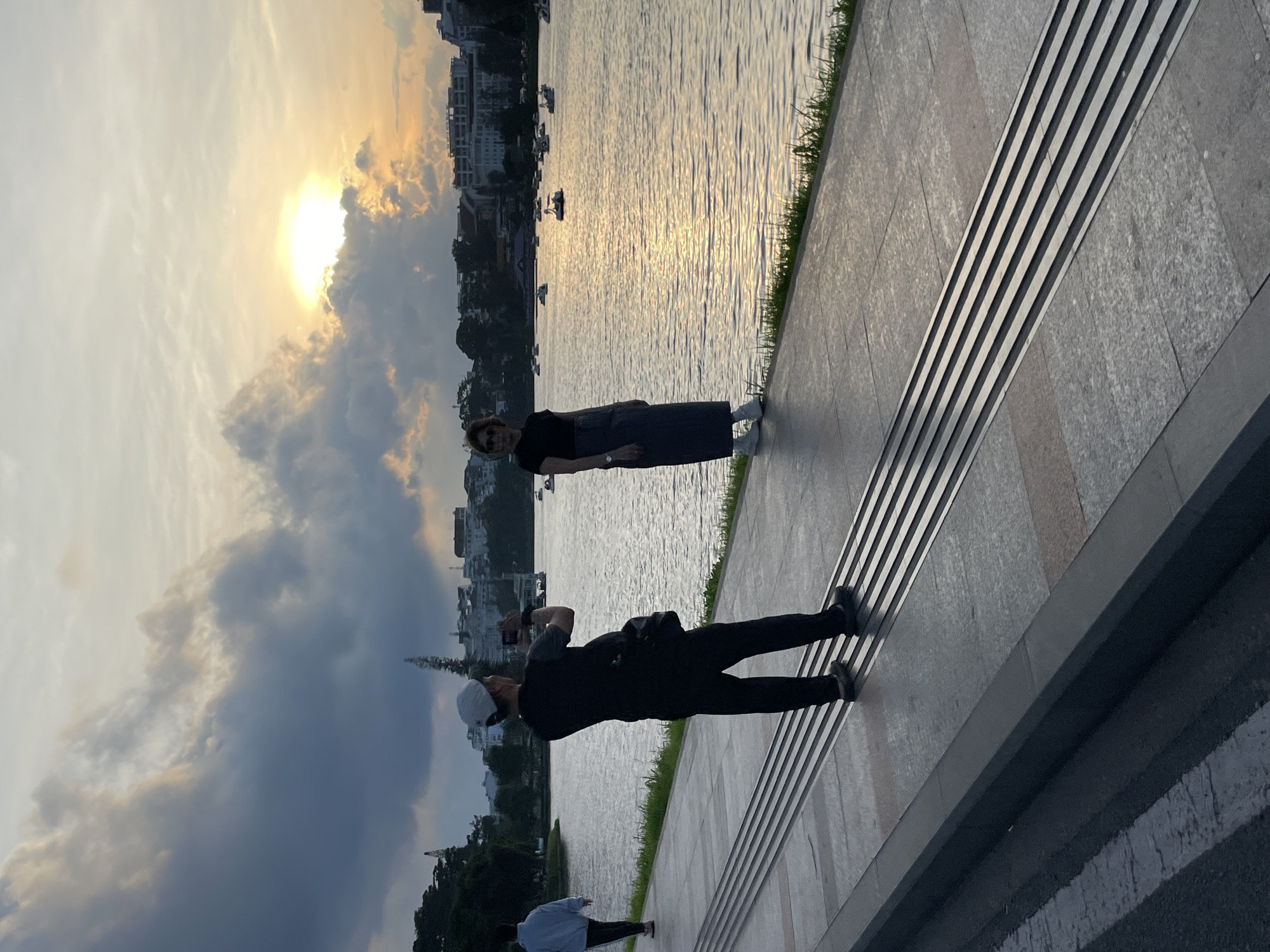 The image depicts a sunset scene along the promenade of Xuan Huong Lake in Da Lat, Vietnam. The sky features a setting sun on the right, emitting a bright golden light partially obscured by clouds. Darker, heavier clouds are visible towards the left of the sky. The lake water reflects the golden hues of the setting sun, creating a shimmering surface dotted with numerous small boats, including several white swan pedal boats, in the mid-ground.
In the foreground, a paved stone promenade runs horizontally, bordered by a narrow strip of green grass adjacent to the lake. A distinct section of tactile paving, consisting of parallel raised lines, is embedded in the center of the promenade. To the far left, a person in a light blue hoodie and dark pants walks away from the camera, heading left along the walkway.
Center-left, a man in a dark t-shirt, dark pants, and a white baseball cap is silhouetted against the bright background. He is turned away from the camera, holding a smartphone horizontally to take a photograph. A black shoulder bag is visible on his left side.
To his right, a woman, also largely silhouetted, stands near the lake's edge. She is wearing a dark t-shirt, a long dark skirt, and white sneakers, with sunglasses on. Her body is slightly angled towards the right, and she appears to be posing for the man's photograph. A watch is visible on her left wrist.
Across the lake, in the background, a cityscape with various buildings and structures is visible, interspersed with trees. The overall lighting suggests late afternoon or early evening, with the low sun casting long shadows and creating strong backlighting.