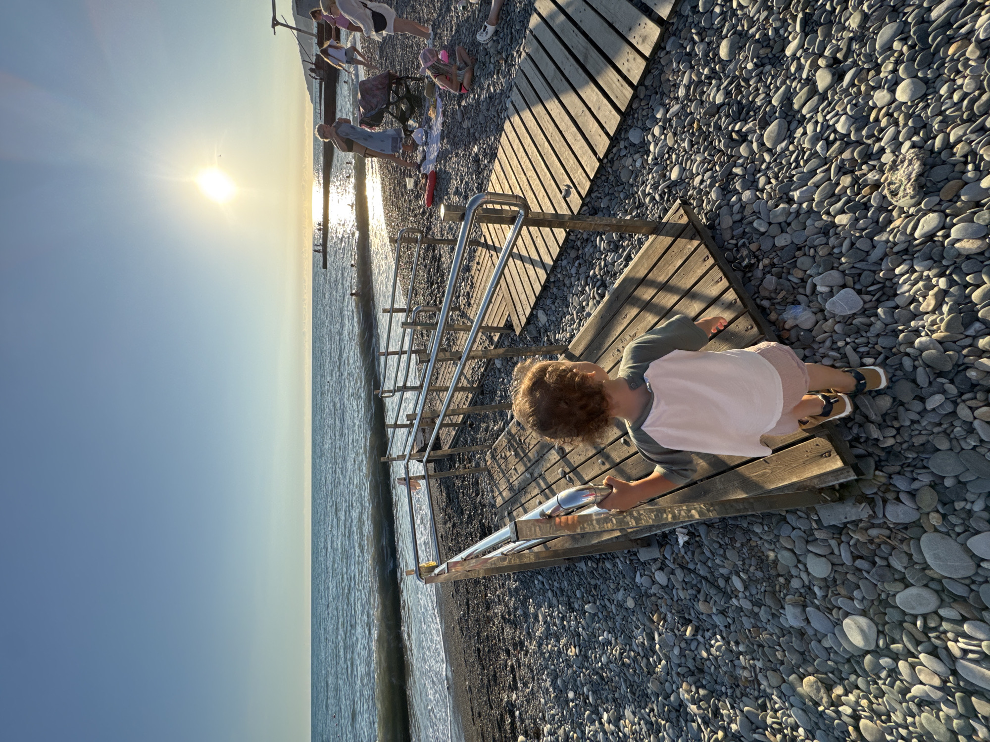 A young child with curly brown hair, wearing a gray and white t-shirt and pink shorts, walks on a wooden ramp on a pebble beach. The ramp has metal railings. In the background, the Black Sea stretches to the horizon under a bright, setting sun. Several people are visible on the beach and in the water. The scene is set on a beach in Sochi, Russia, likely in the late afternoon or early evening, given the low angle of the sun and the warm lighting. The overall atmosphere is one of a relaxed summer day at the seaside.