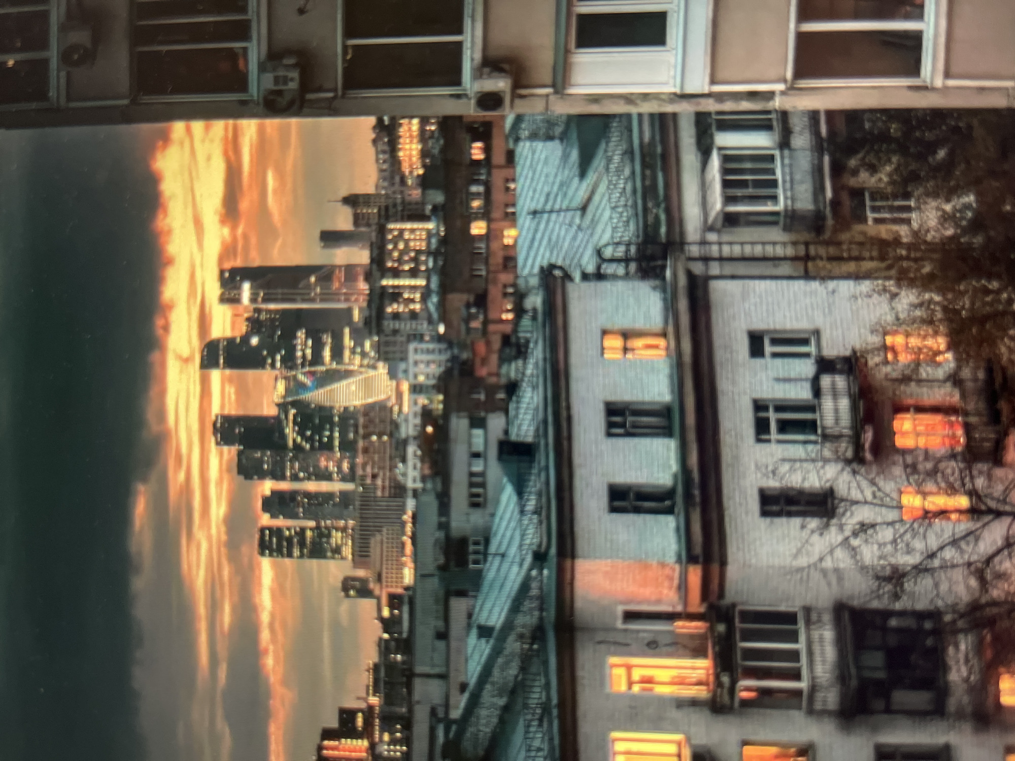 The image depicts a city skyline at dusk. In the foreground, there are multi-story apartment buildings with lit windows, some featuring balconies. The roofs of these buildings are covered in what appears to be metal sheeting. Beyond the apartment buildings, a dense collection of modern skyscrapers rises against a dramatic sky. The sky is a mix of dark, ominous clouds and vibrant orange and yellow hues, suggesting a sunset. The skyscrapers are illuminated with numerous lights, indicating that the city is active. To the far right of the image, the side of another apartment building is visible, also with windows and an air conditioning unit. The overall scene conveys a juxtaposition of older residential architecture with a contemporary business district. While the location is specified as Khimki, Russia, the image itself does not contain any explicit visual cues to confirm this specific location beyond the general appearance of a Russian city. There are no people or animals clearly visible in the image.