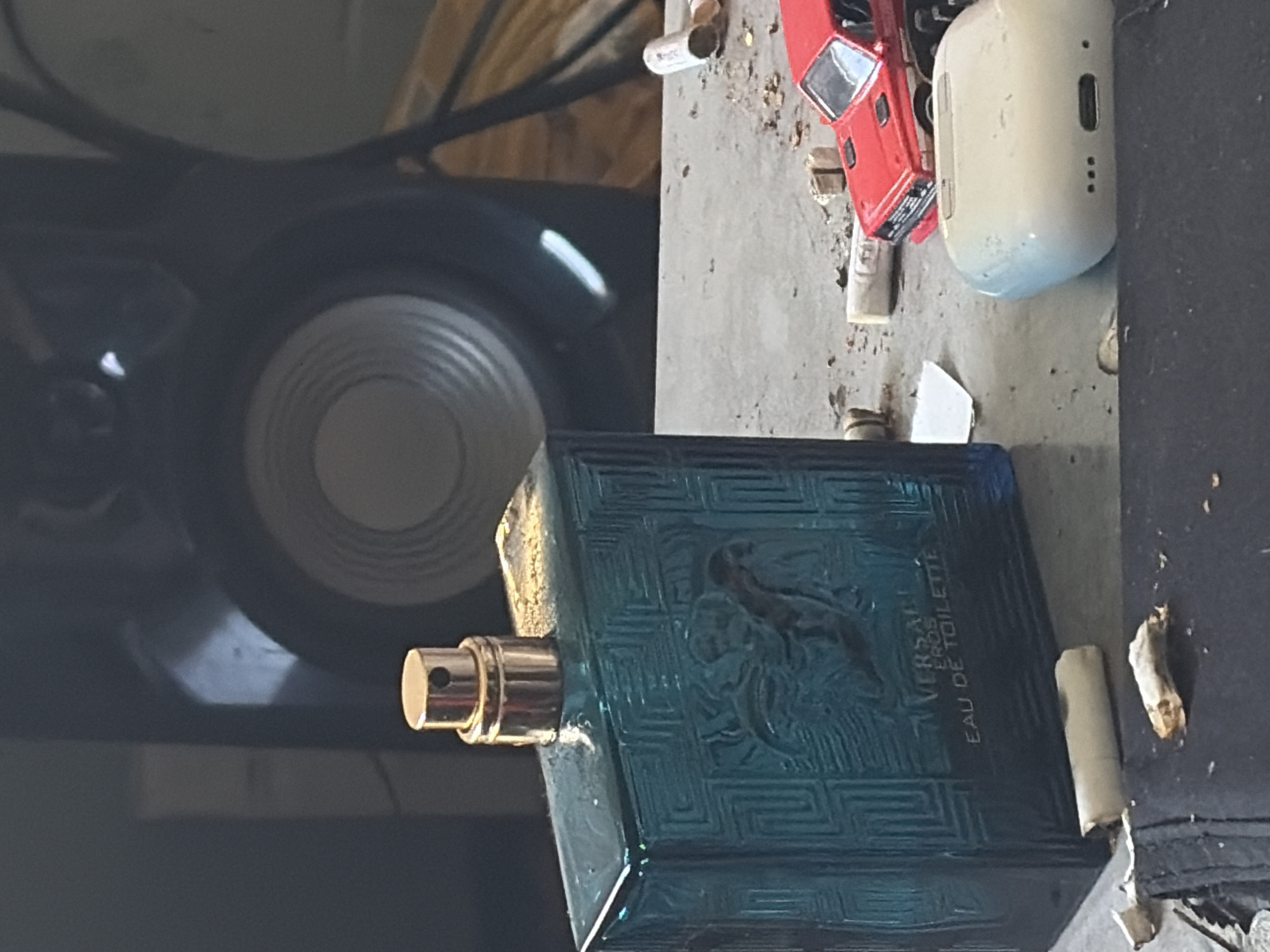A rectangular, dark teal glass bottle of Versace Eros Eau de Toilette perfume lies horizontally across a dirty, textured grey surface. The bottle features a textured Greek key pattern along its edges and a raised relief of a Medusa head on its side. A gold-colored sprayer mechanism protrudes from its left side.
To the upper right of the perfume bottle, a small red toy car is visible, appearing to be a classic sports car model. Above the toy car, a pale, off-white or light blue earbud charging case, resembling an AirPods case, is present. Several discarded cigarette butts or pieces of rolled paper are scattered on the dusty surface, some near the perfume bottle and others further away.
The background on the left side of the image is dark and out of focus, revealing a large, dark circular object with a silver or grey concentric pattern, likely a speaker cone. Behind this, blurry dark shapes and light-colored wooden structures or beams can be discerned. The overall setting appears to be a cluttered and somewhat neglected indoor space, possibly a garage, workshop, or storage area, given the scattered debris and type of objects.
Visible text on the perfume bottle clearly reads "VERSACE EROS EAU DE TOILETTE," with "VERSACE" also visible in smaller print above "EROS." The Medusa head logo is prominent. The entire image appears to be rotated 90 degrees counter-clockwise.