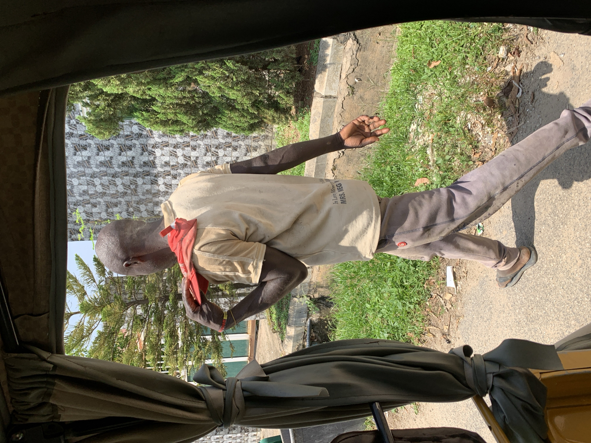 A bald Black man wearing a tan t-shirt and gray pants walks away from the camera outside on a sunny day. He has a red cloth draped over his left shoulder, and his left hand is holding the cloth to his shoulder. He is wearing dark sandals. The man is walking along a paved path beside a grassy area with some trees. Behind him is a building with a patterned facade and some more trees. The image is framed by dark curtains on the right and tan curtains on the left, suggesting the photo was taken from inside a vehicle, possibly an auto-rickshaw. The scene appears to be in Abuja, Nigeria.