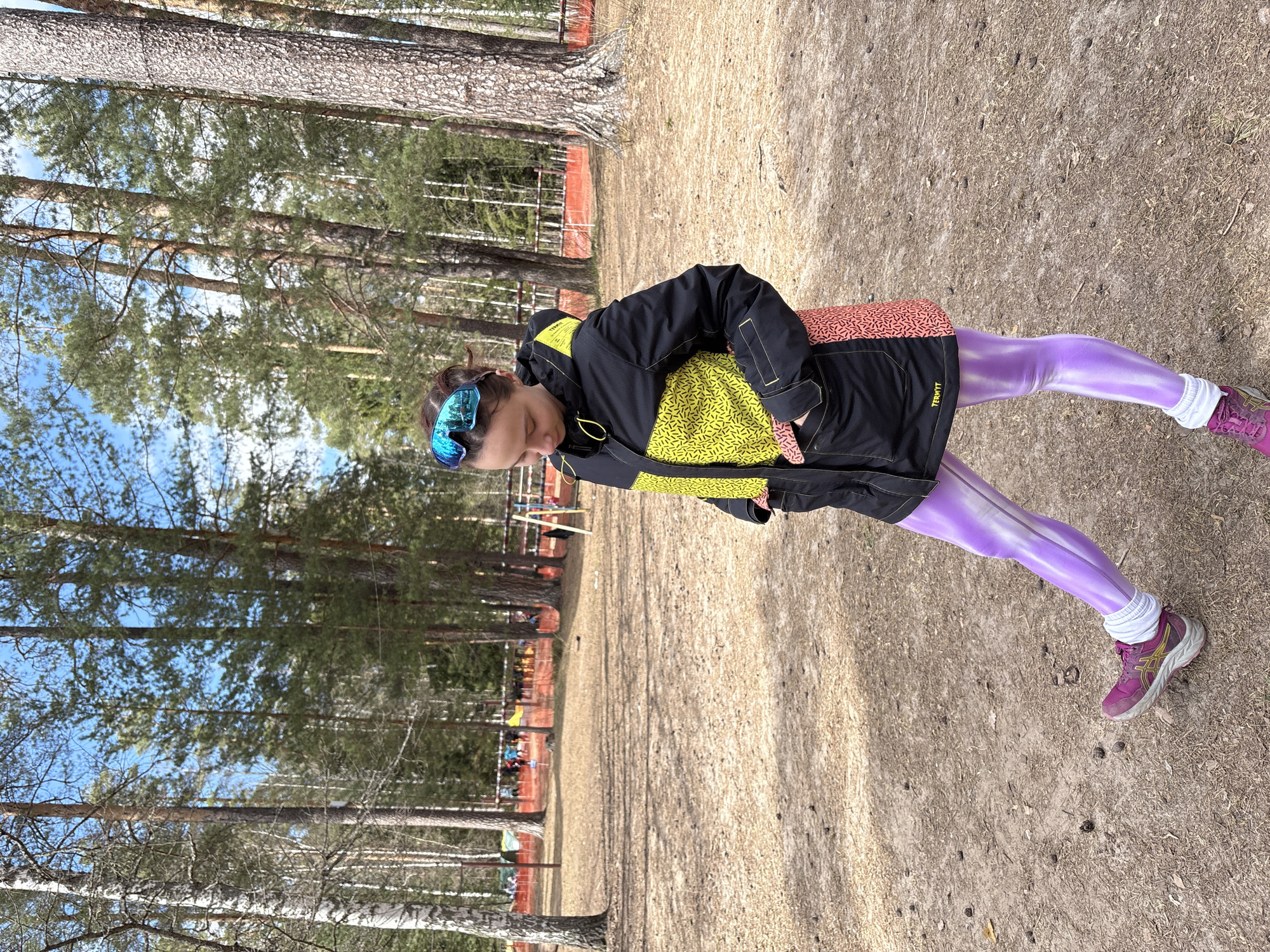 A person is standing on a dirt path in a wooded area. The person is wearing a black jacket with yellow patterns, purple leggings, and purple running shoes with white socks. They also have blue-tinted goggles on their head. The environment consists of tall trees and a dirt ground. The location is Lesnoye, Russia. There appears to be a fenced-off area with people standing behind it in the distance.