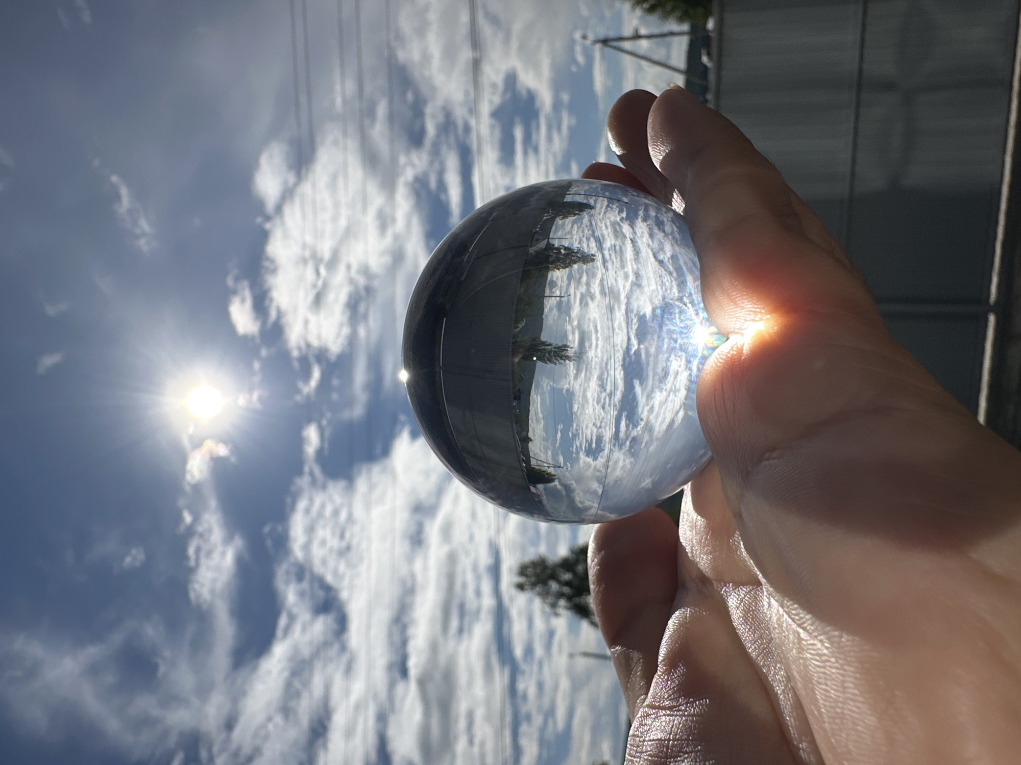 A close-up, low-angle photograph captures a human hand holding a clear, spherical crystal ball outdoors under a bright, partly cloudy sky in the city of Unknown, Unknown. The hand, positioned in the lower-left to central foreground, cradles the crystal ball with its palm facing slightly upward. Strong, direct sunlight illuminates the hand, highlighting skin texture and creating bright reflections. The crystal ball, the primary subject, reflects an inverted image of the surrounding environment, showing white clouds and a blue sky at the bottom, and dark, inverted tree lines and horizontal power lines at the top. A concentrated, bright light flare, likely from the sun, is visible at the bottom center of the crystal ball and reflects onto the palm. In the blurred background, a bright sun emits visible rays from the upper-right quadrant. The sky is predominantly blue with numerous scattered white cumulus clouds. Multiple dark, thin horizontal lines, identified as power lines, stretch across the middle ground. Out-of-focus dark green trees are visible in the mid-ground, with one prominent silhouette to the left of the crystal ball. Fences and parts of obscured structures are faintly visible in the lower-right background.