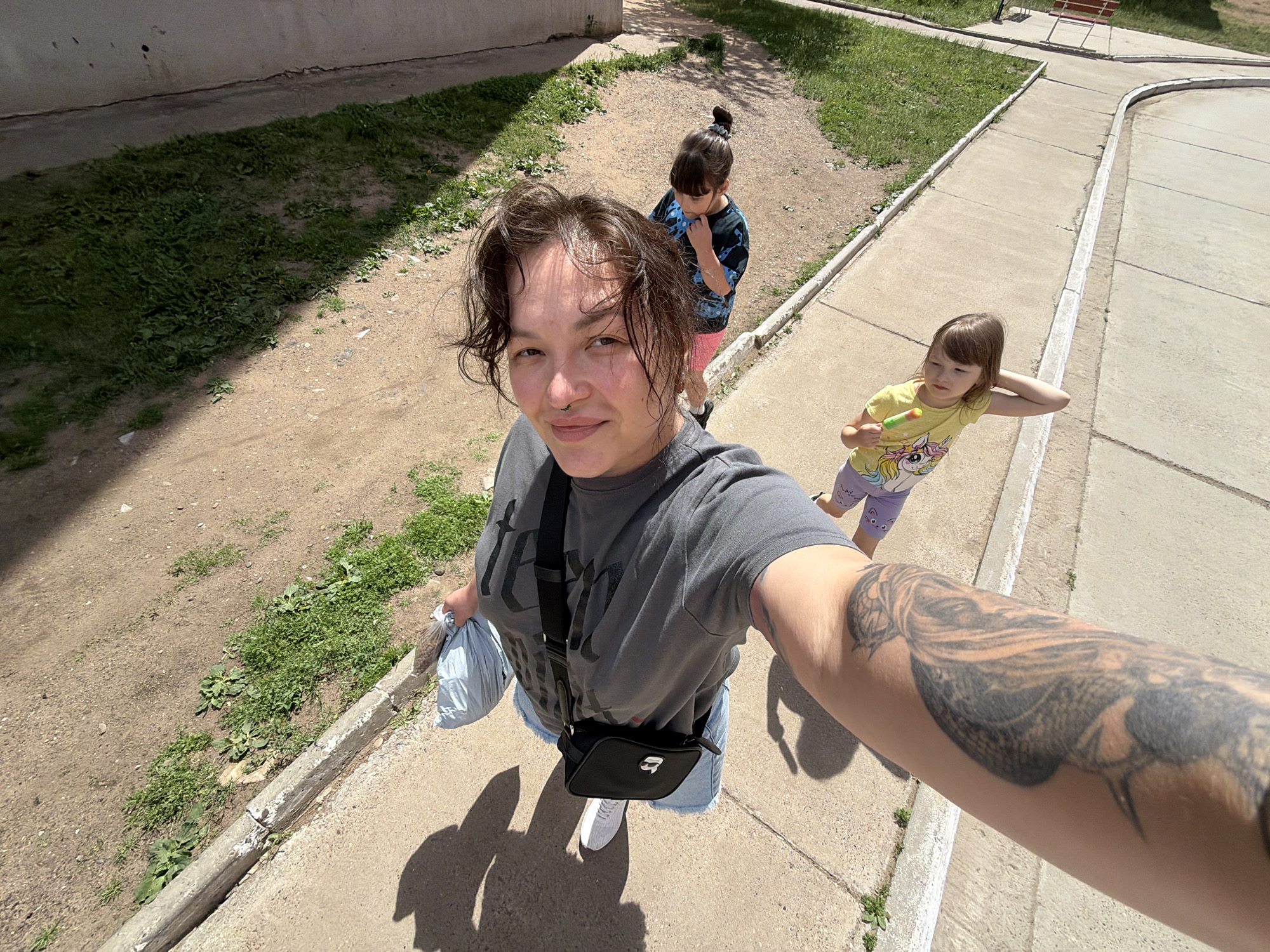 A woman with a septum piercing and a large tattoo on her arm is taking a selfie outdoors. She is wearing a gray t-shirt, denim shorts, and white sneakers, and carrying a black crossbody bag and a light blue plastic bag. Two young girls are visible in the background. One girl, wearing a black and blue tie-dye shirt and pink pants, stands behind the woman, looking down. The other girl, wearing a yellow t-shirt with a unicorn graphic and purple pants, is to the woman's right, holding a popsicle and looking away. They are on a concrete pathway next to a dirt and grass area. The scene appears to be in a residential area, possibly a courtyard in the city of Chaikovsky, Russia, with a concrete wall and some sparse vegetation visible.