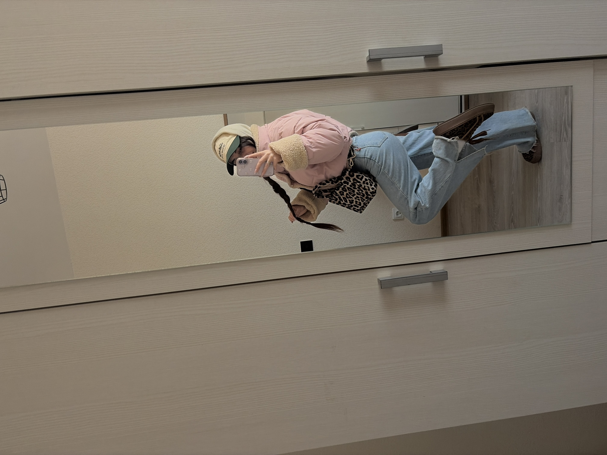 A mirror reflects a woman posing in a room. The woman wears a beige baseball cap, a pink jacket with a beige fleece collar, light blue jeans, and brown shoes. She holds a light purple smartphone, presumably taking a picture of herself. A leopard print bag hangs from her side. Her long, dark braid is visible. The mirror is set into a light beige, wooden wardrobe, with silver handles on the doors. The wall behind the woman is beige. The floor in the reflection is wood-toned. The location is likely inside a private residence. There are no indications that the photograph was taken in Saint Petersburg, Russia.