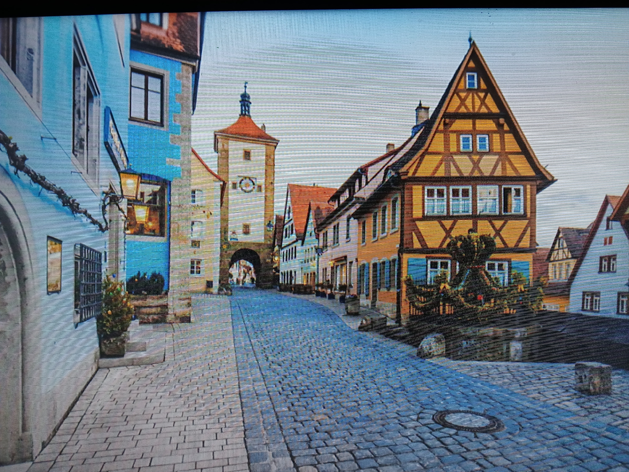 The image depicts a street scene in what appears to be a European town, not Juan Griego, Venezuela. A cobblestone street leads towards an arched gateway and a clock tower. The architecture consists of colorful buildings, with distinctive half-timbered houses to the right. There are no visible people or signs of activity, creating a quiet and somewhat historical atmosphere. A small potted tree is visible on the left. The overall impression is of a well-preserved, historic urban landscape.